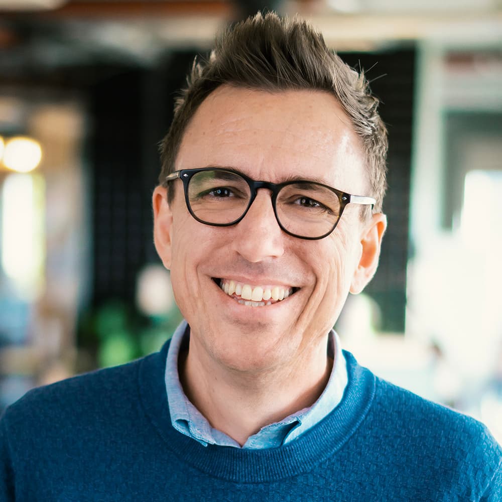 Lächelnde Person mit kurzen Haaren und Brille, die einen blauen Pullover trägt, steht in einem Innenraum mit unscharfem Hintergrund.