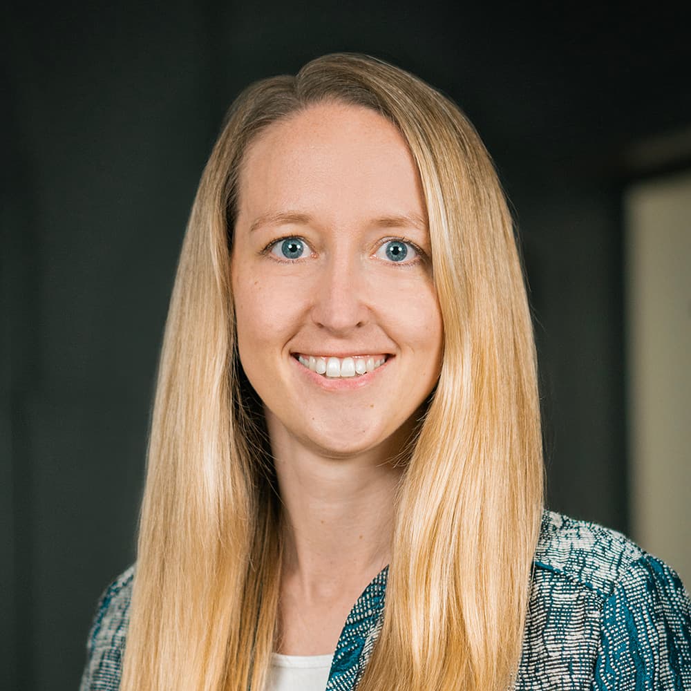 Lächelnde Frau mit langen blonden Haaren, die einen gemusterten Blazer über einem weißen Oberteil trägt, vor einem dunklen Hintergrund.