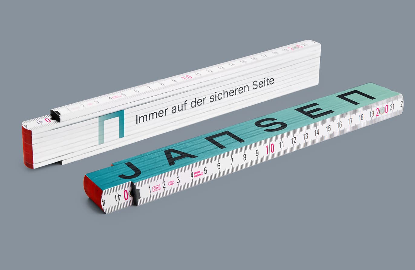 Two folding rulers on a gray background, one blue with "JANSEN" and one white with German text "Immer auf der sicheren Seite."