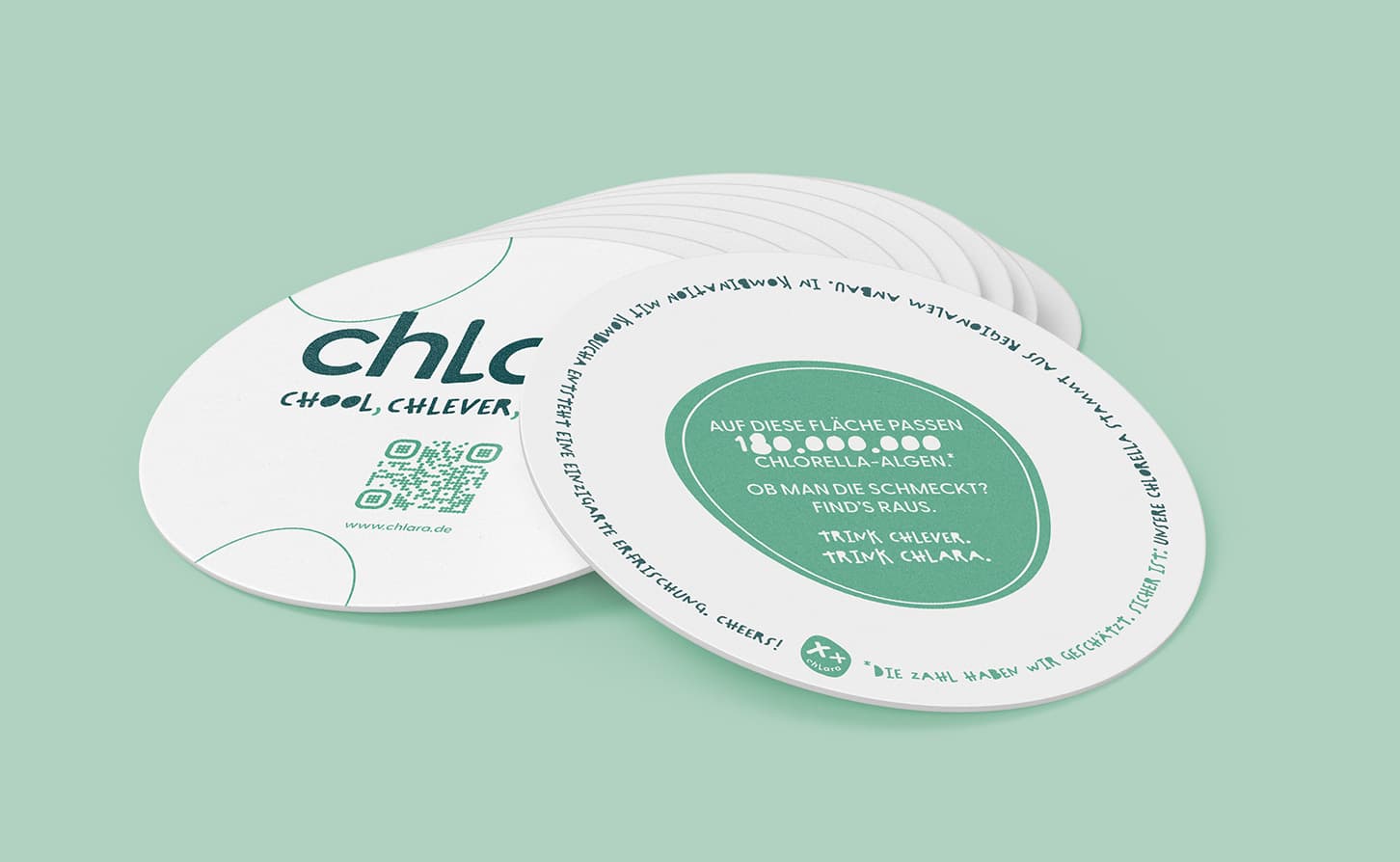 Stapel von runden Werbeuntersetzern der Marke 'chlara', bedruckt mit grüner Schrift und einem QR-Code, auf mintgrünem Hintergrund. Einer der Untersetzer zeigt die Information, dass 180.000.000 Chlorella-Algen auf diese Fläche passen, mit dem Aufruf: 'Trink Chlever. Trink Chlara.'