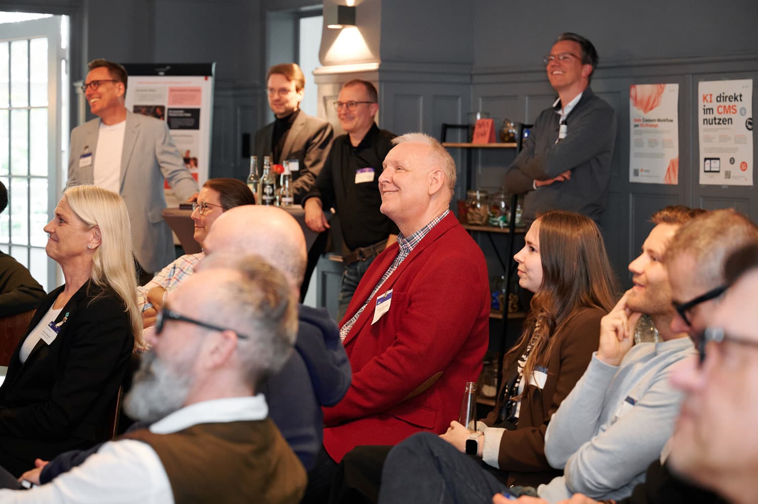 Mehrere Teilnehmende sitzen in einem Veranstaltungsraum und schauen aufmerksam nach vorn; im Hintergrund stehen weitere Personen an Stehtischen, an der Wand hängen Infoposter.