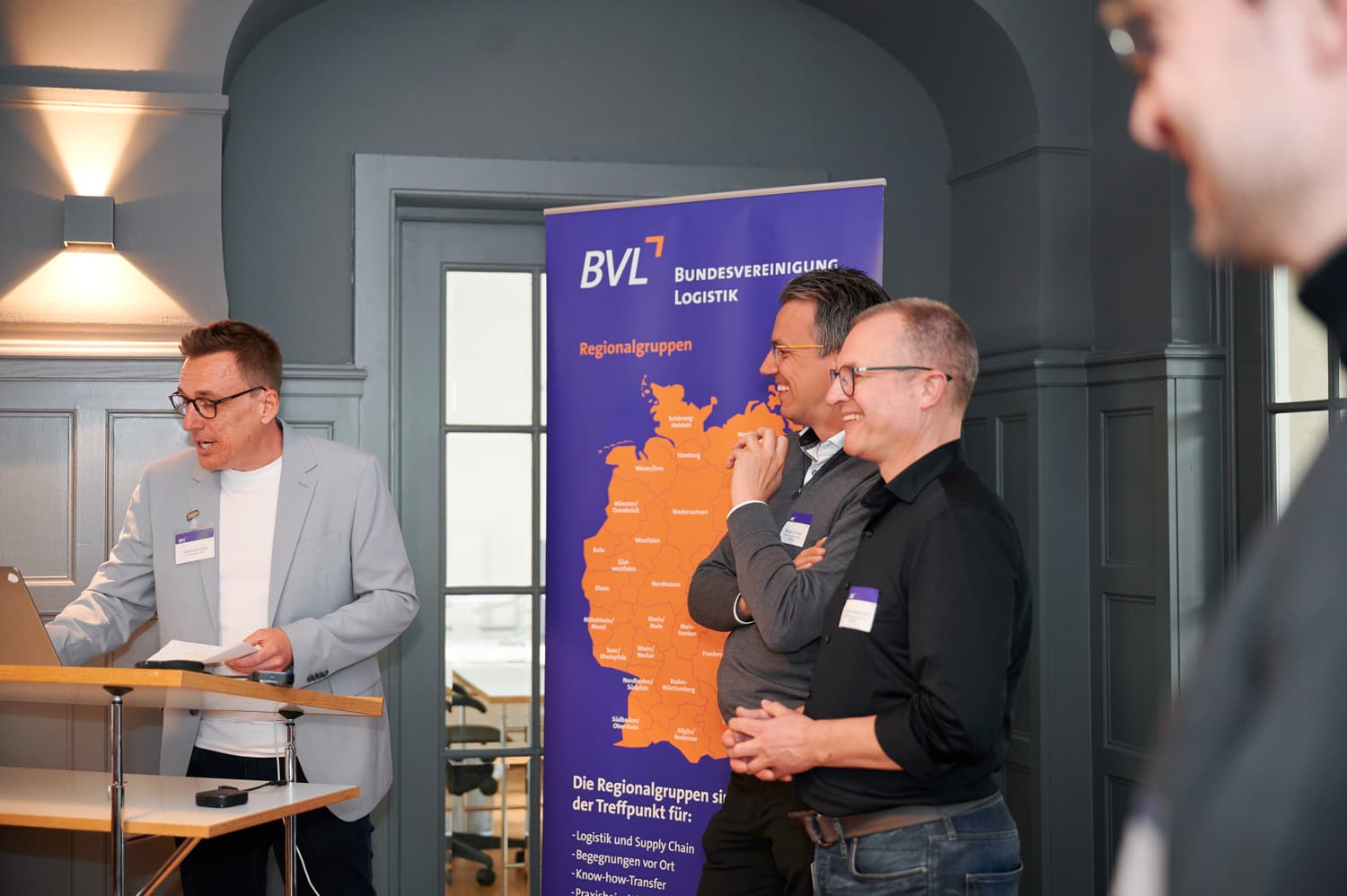 Eine Gruppe von Männern steht bei einer Präsentation vor einer BVL-Logistik-Tafel. Der Präsentierende spricht und die anderen hören lächend zu.