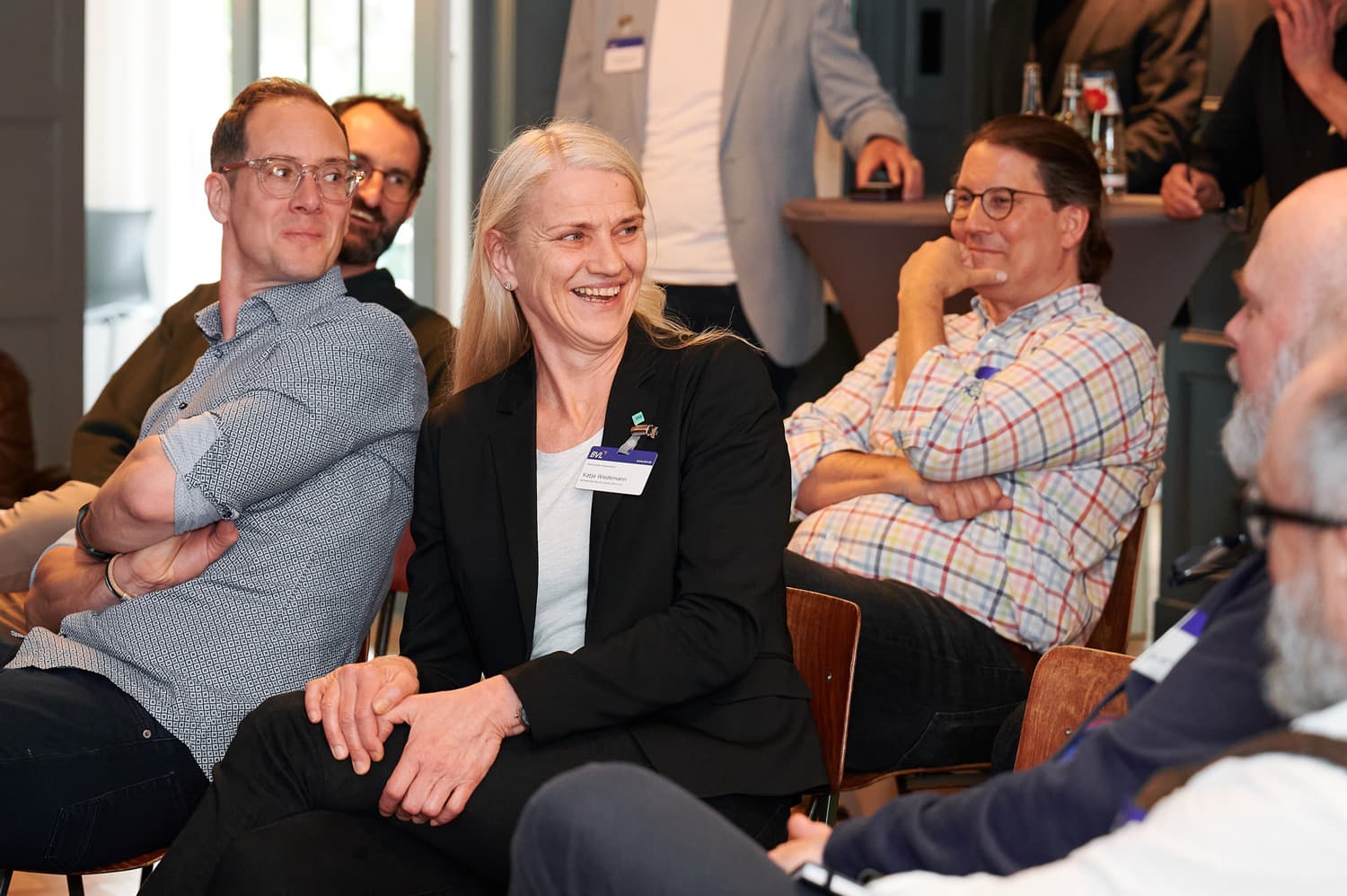 Eine Frau im schwarzen Blazer sitzt in einer Publikumsrunde und schaut lächelnd nach hinten; neben ihr sitzen weitere Teilnehmende, die aufmerksam zuhören.