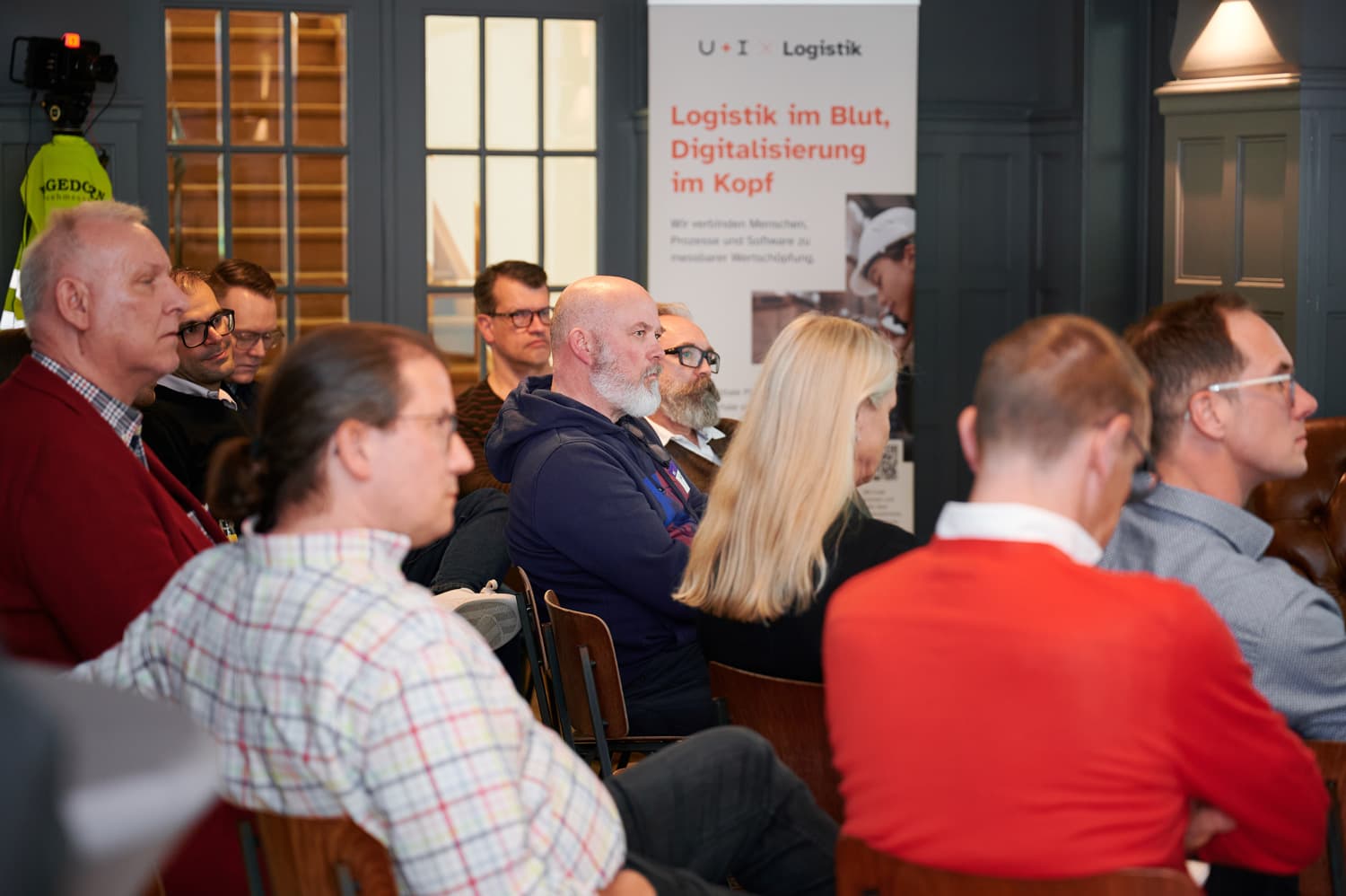 In einem größeren Raum sitzen viele Teilnehmende in Reihen und schauen nach vorn; im Hintergrund hängen Präsentationsbanner und weiteres Infomaterial.