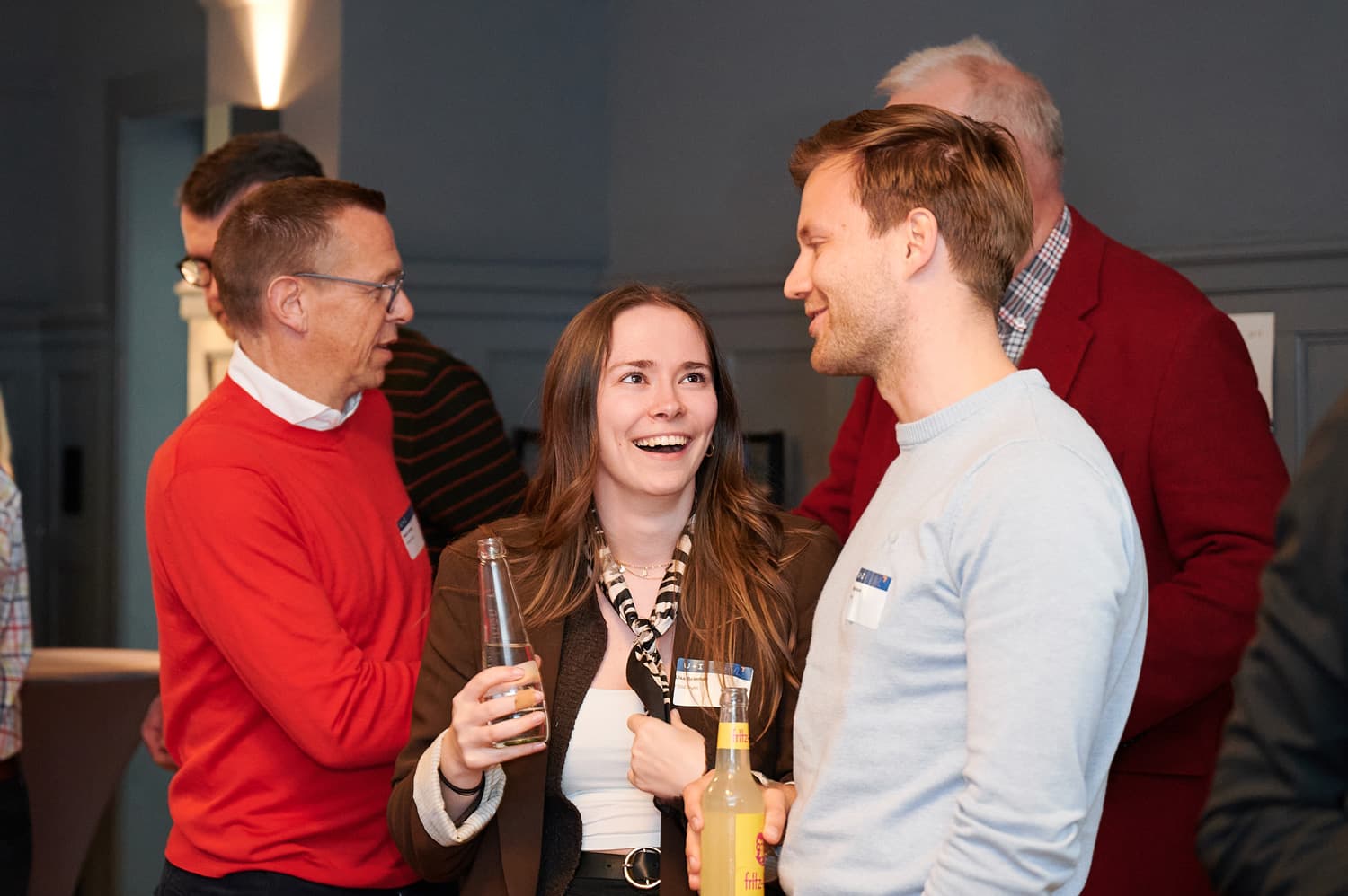 Drei Personen bei einer Veranstaltung im Innenraum, im Hintergrund sind weitere Gäste zu sehen; mehrere halten Getränke in der Hand, alle wirken in ein Gespräch vertieft.