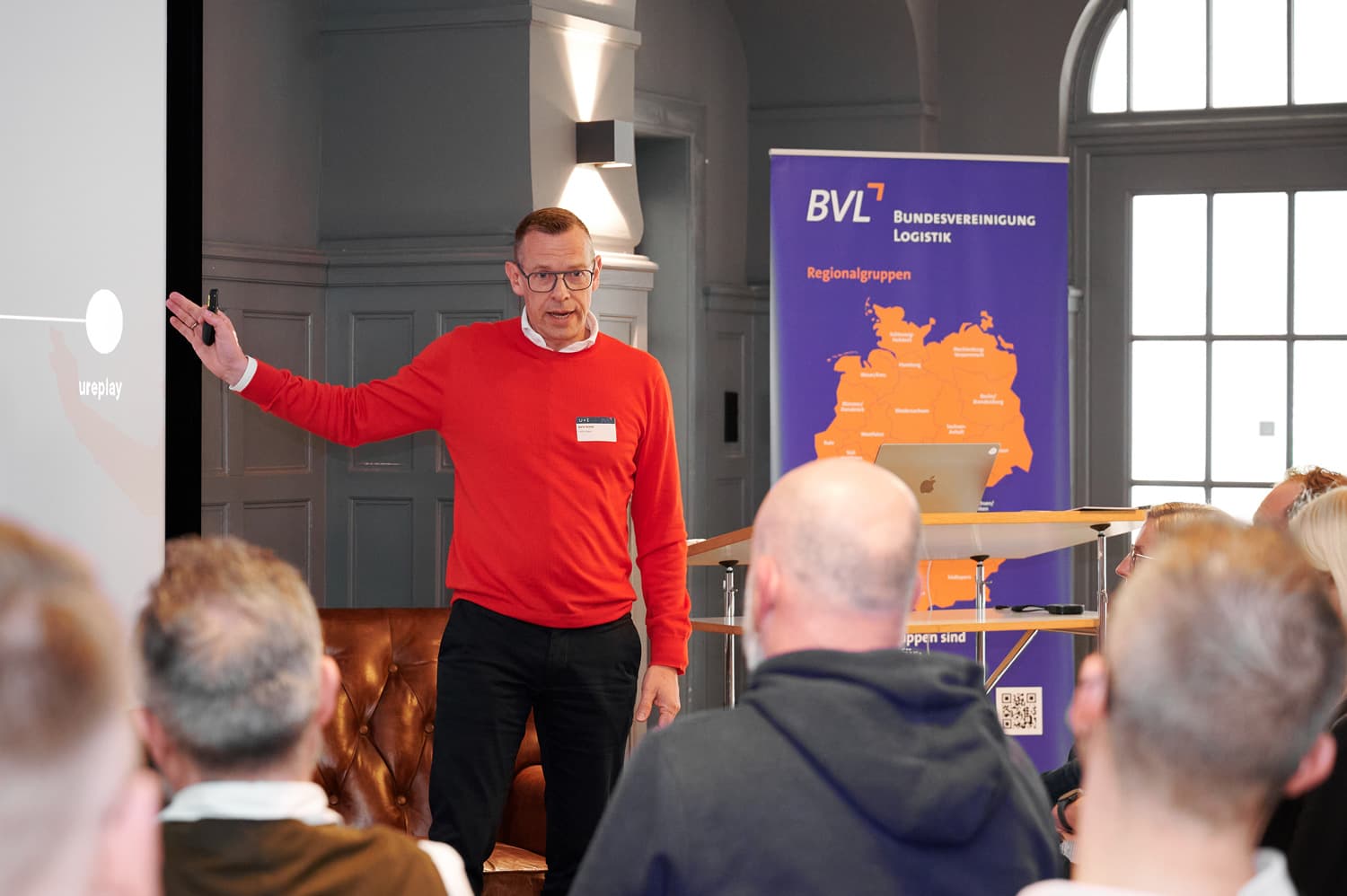 Vortragssituation in einem Saal mit mehreren Personen im Publikum; ein Redner steht seitlich neben der Projektionsfläche und zeigt auf etwas mit seiner Hand.