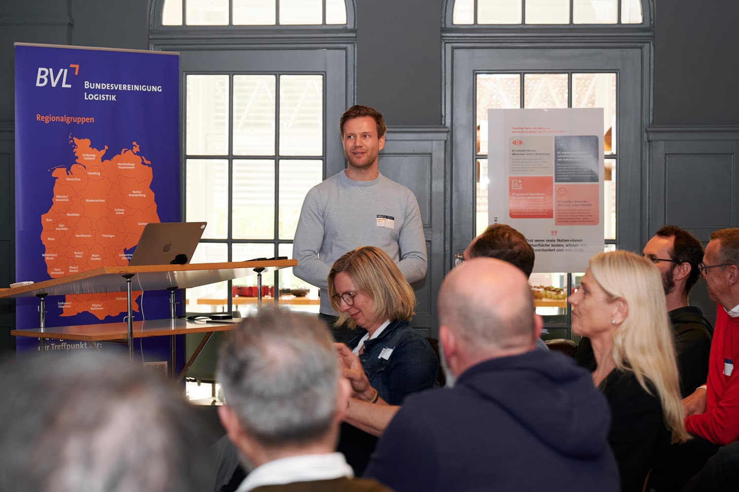 Ein Redner steht vor einer BVL-Karte und spricht vor Publikum; mehrere Zuhörer sind im Vordergrund angeschnitten.