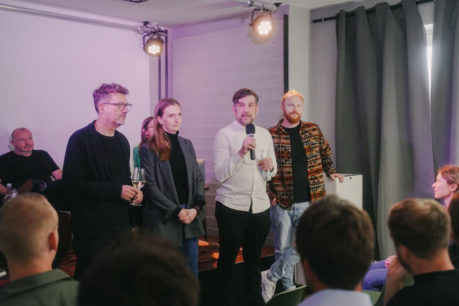 Vier Personen stehen vor einem Publikum in einem kleinen Raum und sprechen, während einer von ihnen ein Mikrofon hält. Die Szene ist in warmes, violettes Licht getaucht. Im Hintergrund sitzen weitere Personen, die der Präsentation aufmerksam folgen. Die Atmosphäre wirkt entspannt und professionell.