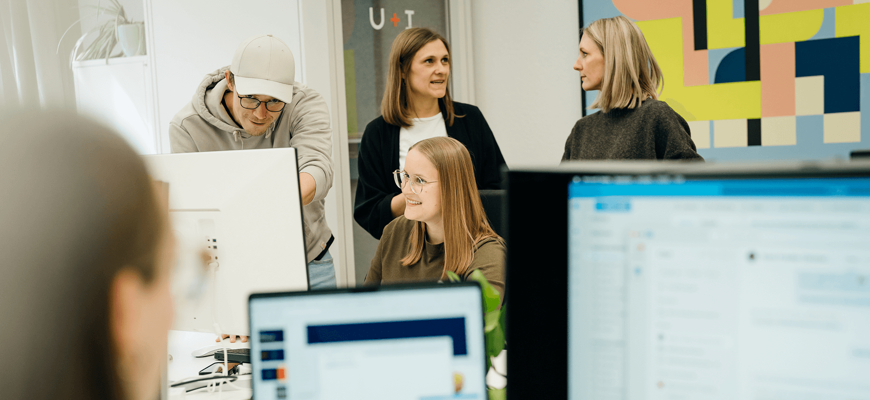 Vier Personen in einem modernen Büro, die sich unterhalten und am Computer arbeiten. Im Hintergrund ist ein buntes abstraktes Gemälde an der Wand zu sehen.