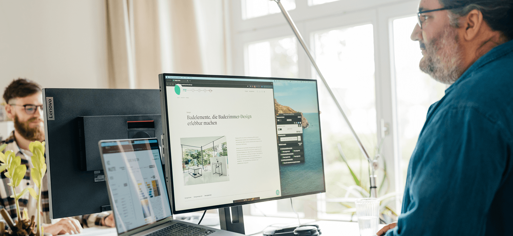 Zwei Männer arbeiten gegenüber voneinander an Schreibtischen mit zwei Monitoren. Ein Bildschirm zeigt das Design einer Website. Sie sitzen in einem hellen Raum mit großen Fenstern und natürlichem Licht.