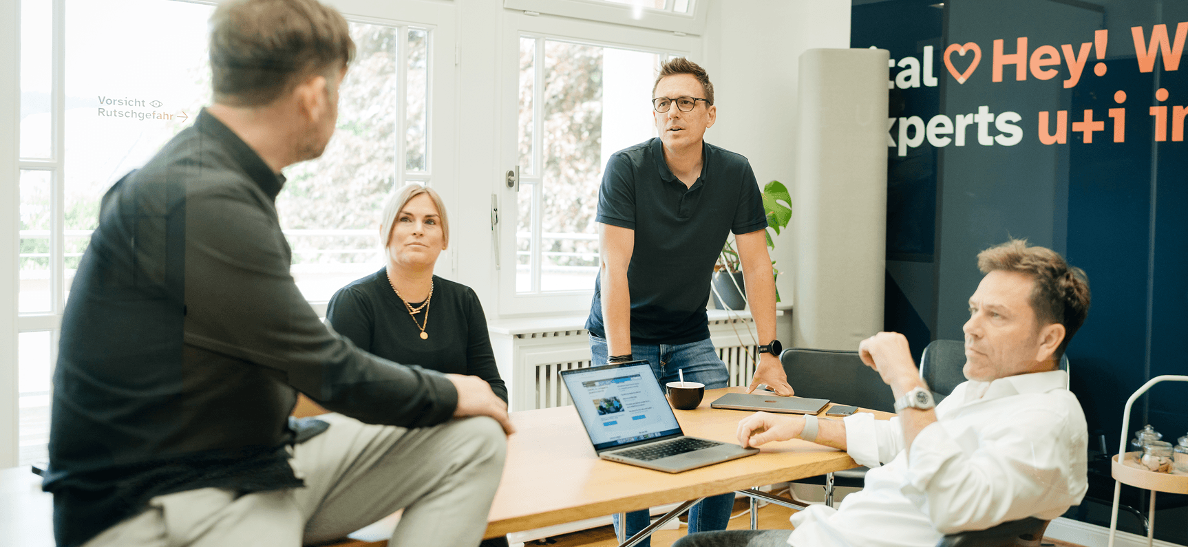 Vier Personen bei einer Diskussion in einem Büro, mit einem Laptop auf dem Tisch. Im Hintergrund sind große Fenster und eine Pflanze zu sehen.