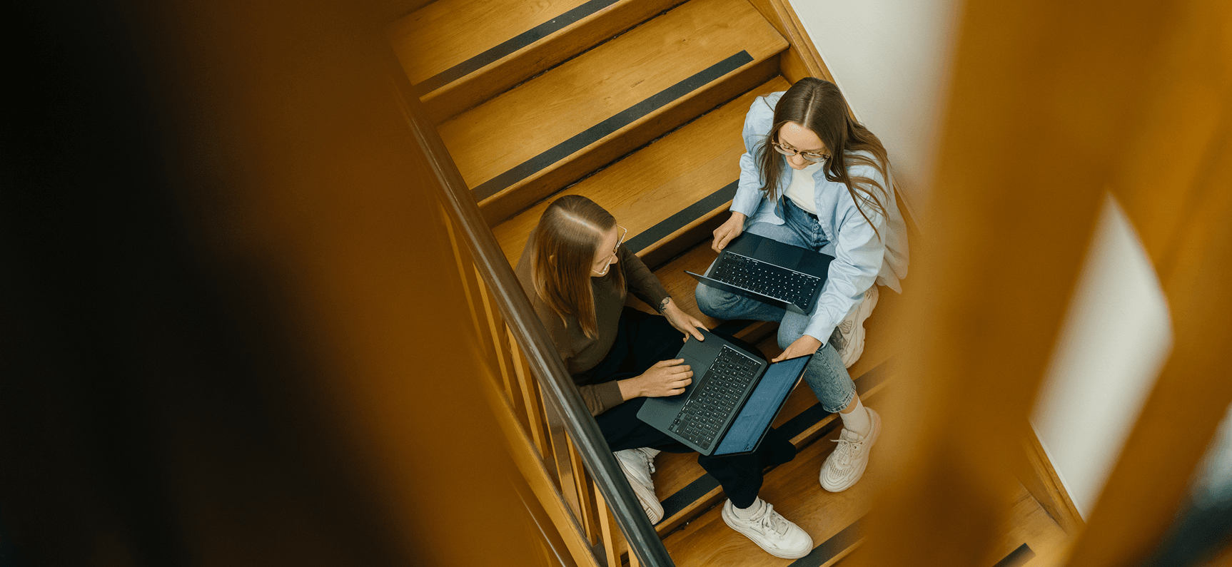 Zwei Personen, die auf einer Holztreppe sitzen und jeweils einen Laptop benutzen. Sie scheinen eine Diskussion zu führen oder zusammenzuarbeiten.