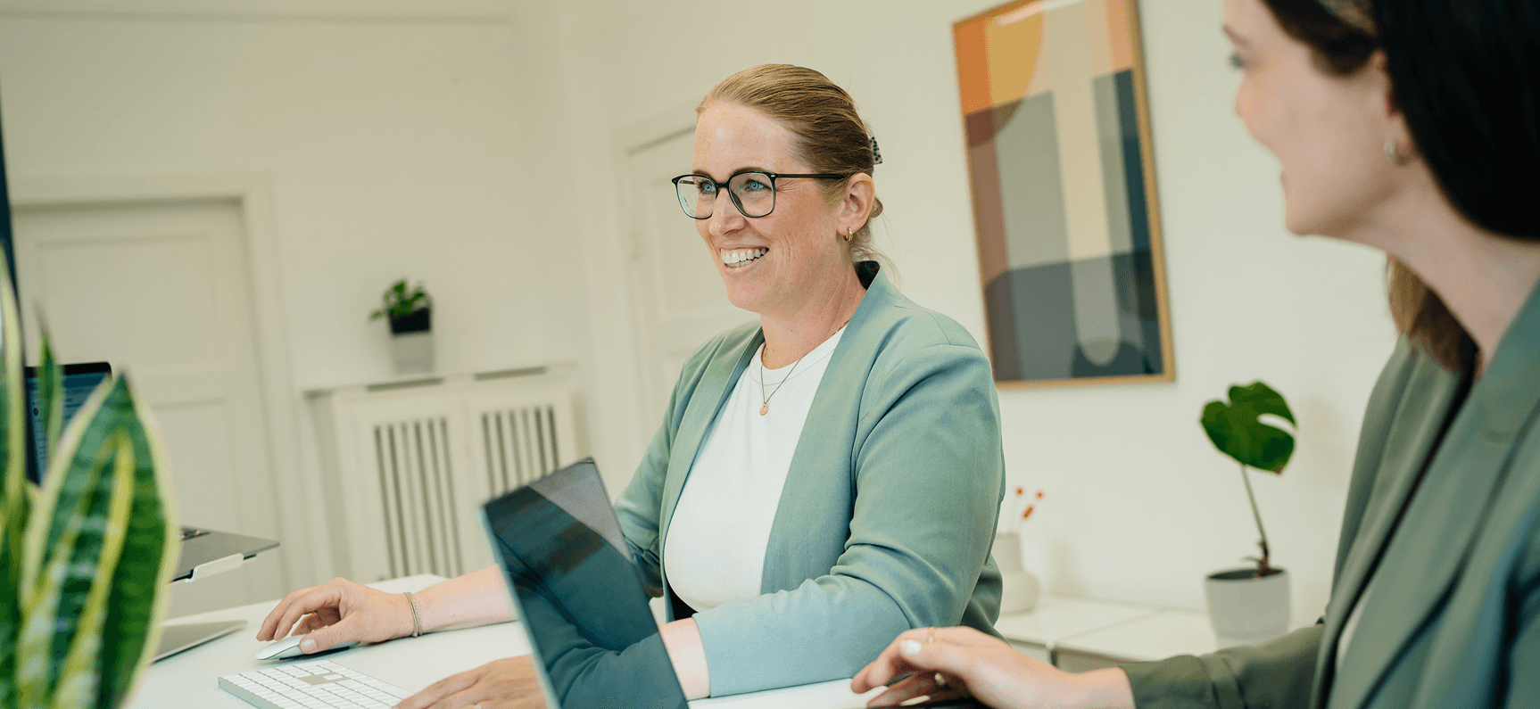 Zwei Personen in einem Büro, eine lächelt in einen Computerbildschirm. Beide tragen grüne Blazer. Im Hintergrund sind eine Pflanze und abstrakte Kunst zu sehen.