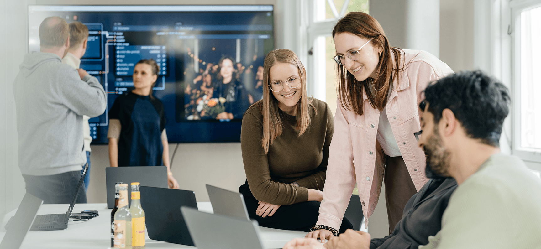 Eine Gruppe von Personen in einem Besprechungsraum, die an ihren Laptops arbeiten und diskutieren, während im Hintergrund ein großer Bildschirm, vor denen drei weitere Personen stehen, zu sehen ist.