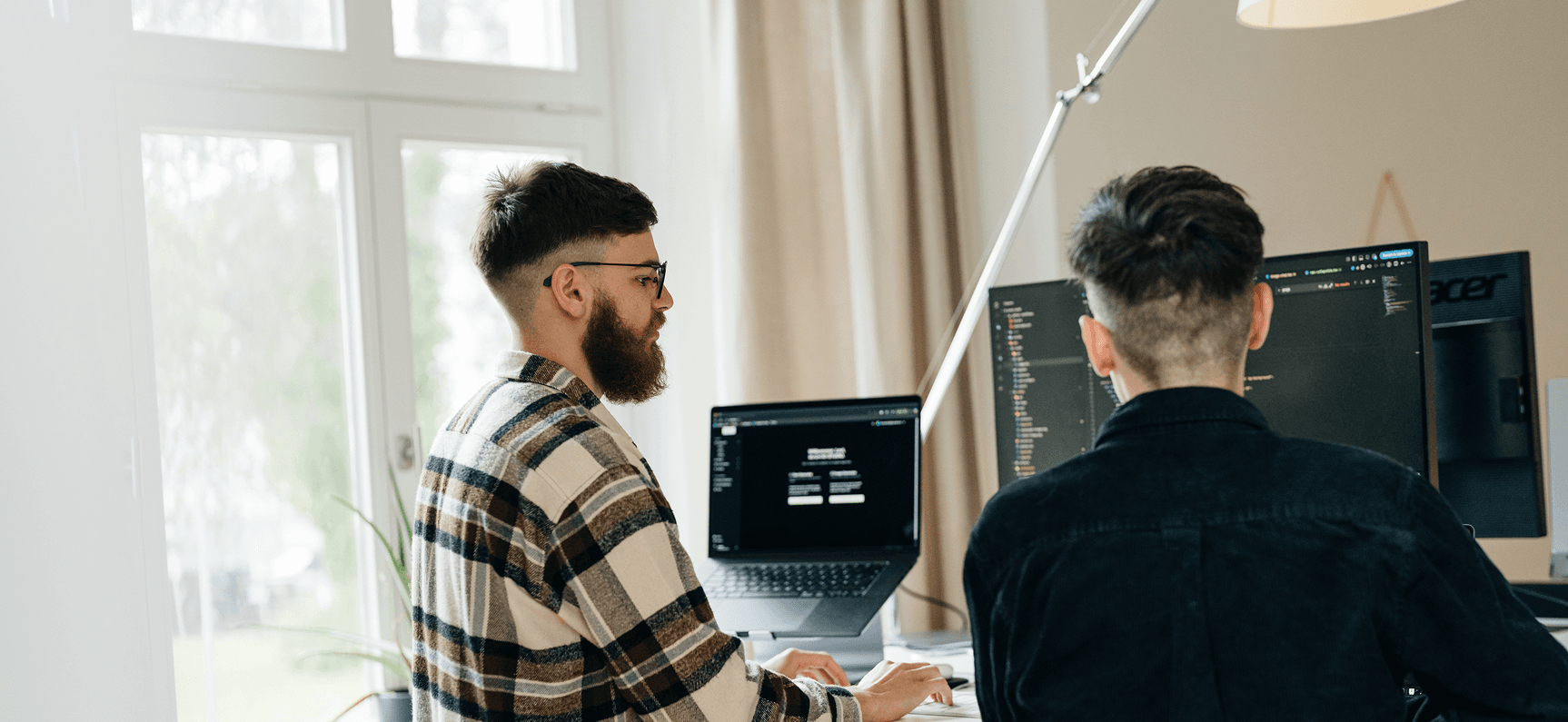 Zwei Personen arbeiten an Computern an einem Schreibtisch, auf den Bildschirmen ist Code zu sehen. Im Hintergrund sind ein Fenster und eine Lampe zu sehen.
