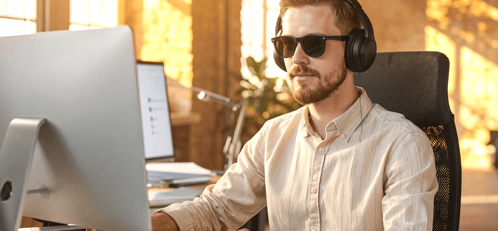 Mann mit Sonnenbrille und Kopfhörern arbeitet an einem Computer in einem sonnendurchfluteten Büro.