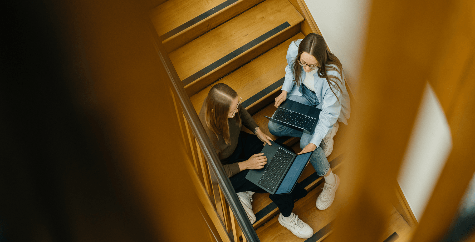 Zwei Personen, die auf einer Holztreppe sitzen und jeweils einen Laptop benutzen. Sie scheinen eine Diskussion zu führen oder zusammenzuarbeiten.