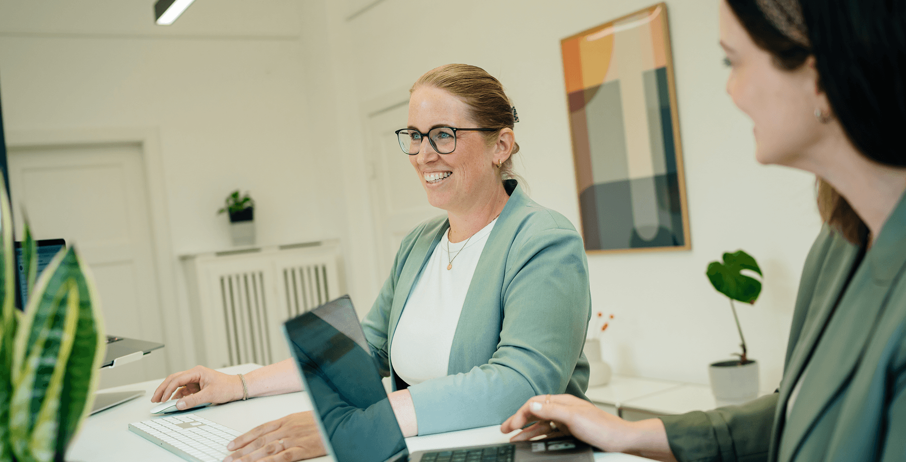 Zwei Personen in einem Büro, eine lächelt in einen Computerbildschirm. Beide tragen grüne Blazer. Im Hintergrund sind eine Pflanze und abstrakte Kunst zu sehen.