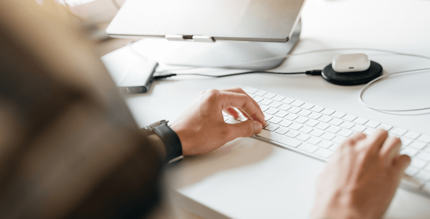 Man sieht die Hände einer Person, die an einem Schreibtisch auf einer weißen Tastatur tippt. Ein Laptop und kabellose Kopfhörer befinden sich auf dem selben Tisch.
