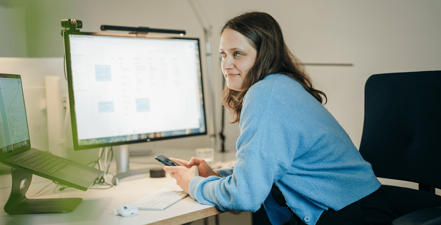 Eine Frau in einem blauen Pullover sitzt an einem Schreibtisch und hält ein Telefon in der Hand, während im Hintergrund große Computermonitore Termine anzeigen.