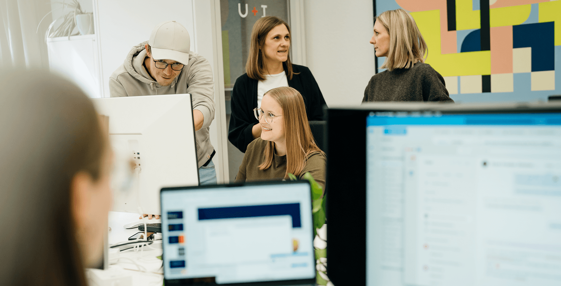 Vier Personen in einem modernen Büro, die sich unterhalten und am Computer arbeiten. Im Hintergrund ist ein buntes abstraktes Gemälde an der Wand zu sehen.