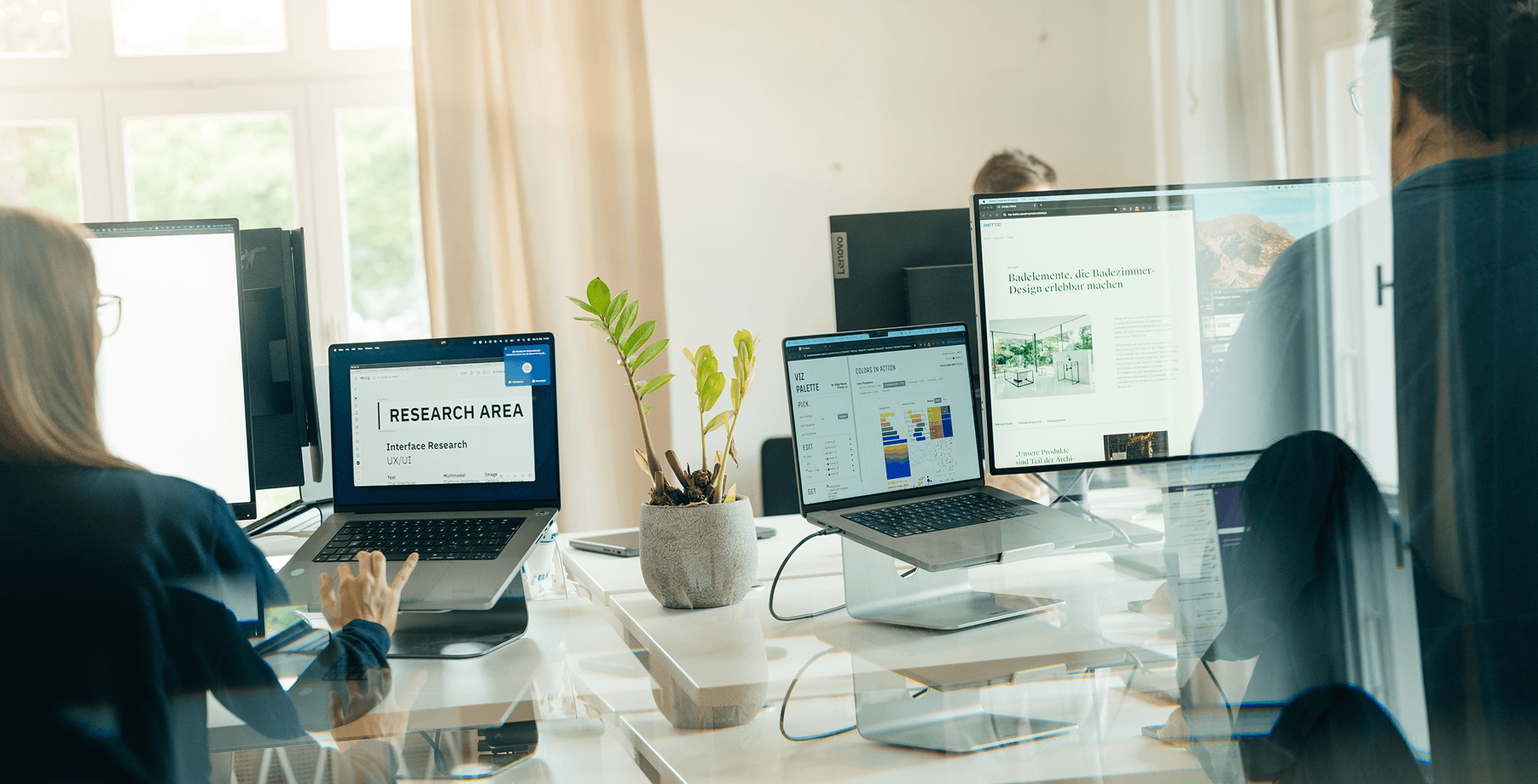Menschen arbeiten in einem Büro mit Laptops und Monitoren, umgeben von natürlichem Licht und einer Topfpflanze.