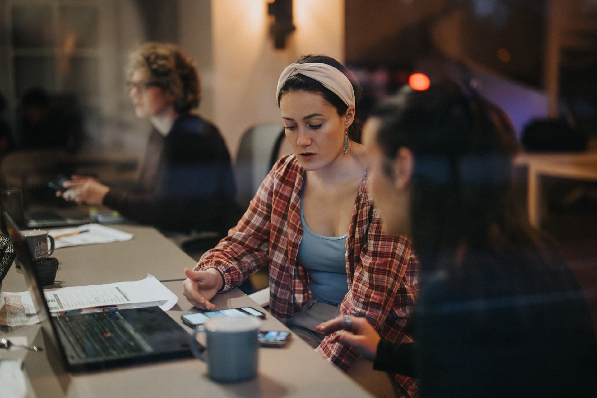 Zwei weibliche Kolleg+innen sprechen an einem Büro-Arbeitsplatz über ein Thema und schauen dabei auf technische Smartphones.