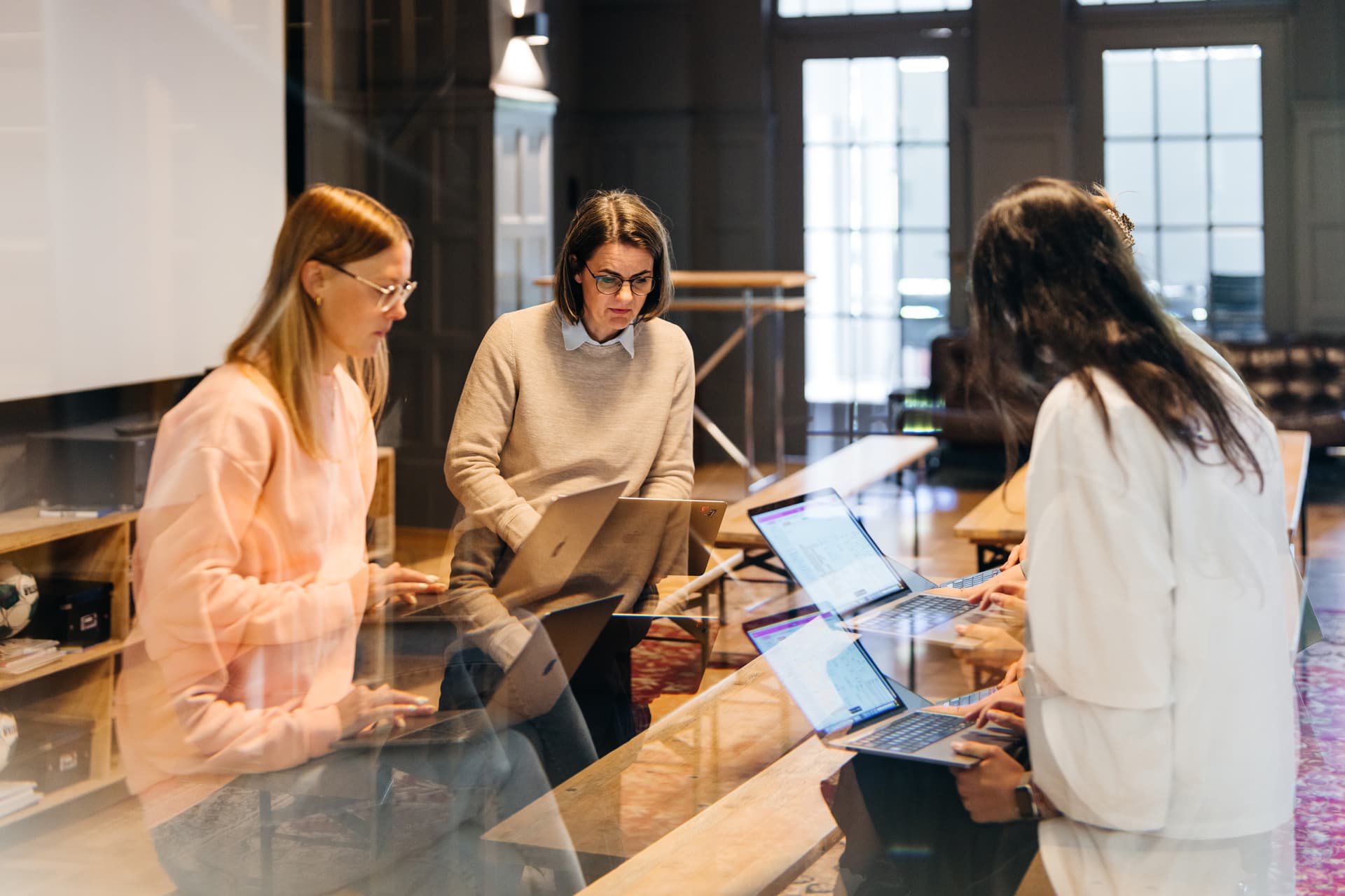 Drei Personen sitzen in einem modernen Besprechungsraum in einer gemütlichen Atmosphäre, arbeiten konzentriert an ihren Laptops und tauschen sich aus. Die Szene ist durch eine Glasscheibe fotografiert, was leichte Spiegelungen erzeugt.