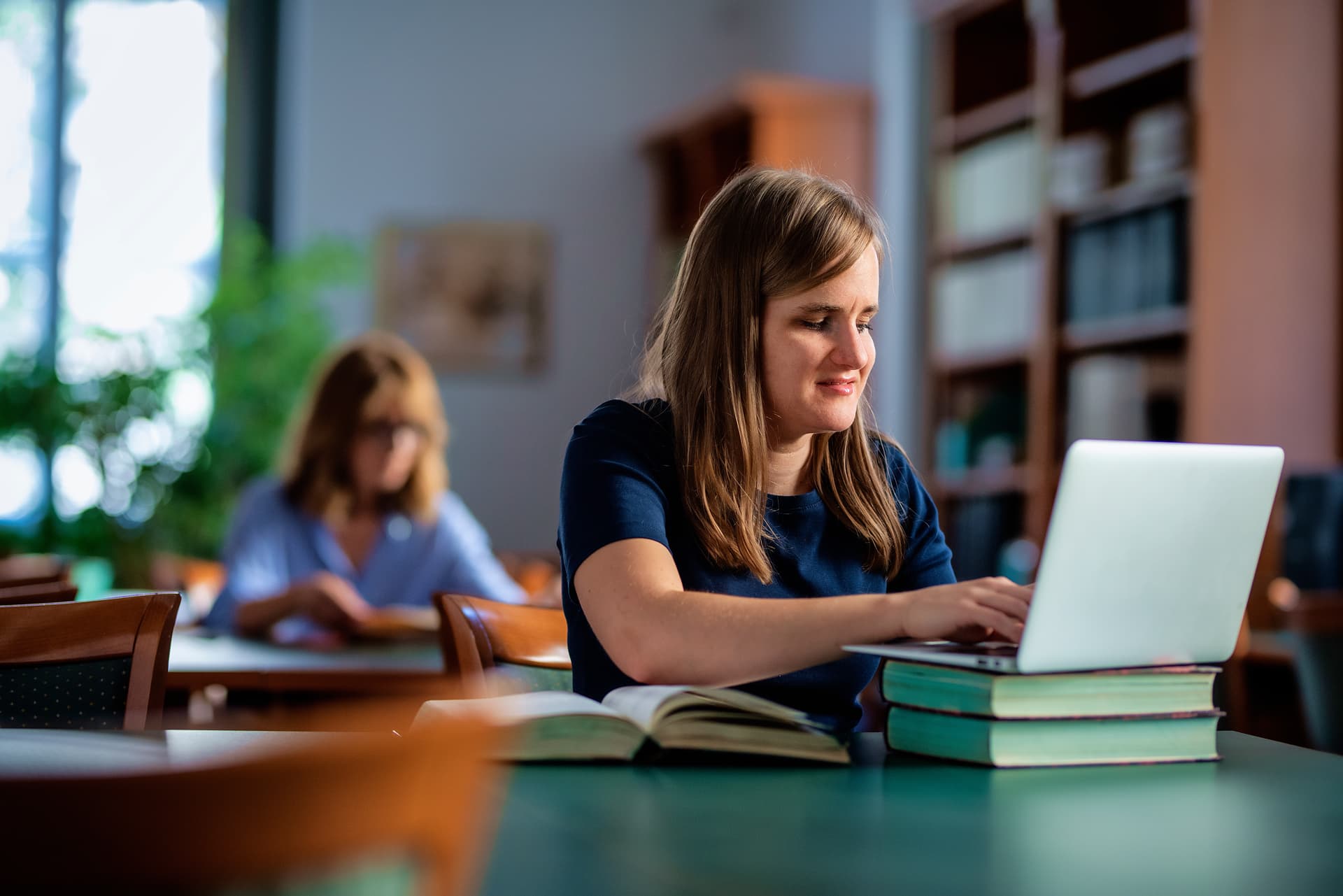 Eine Frau mit Sehbehinderung sitzt in einer Bibliothek an einem Tisch und recherchiert etwas in ihrem Laptop.