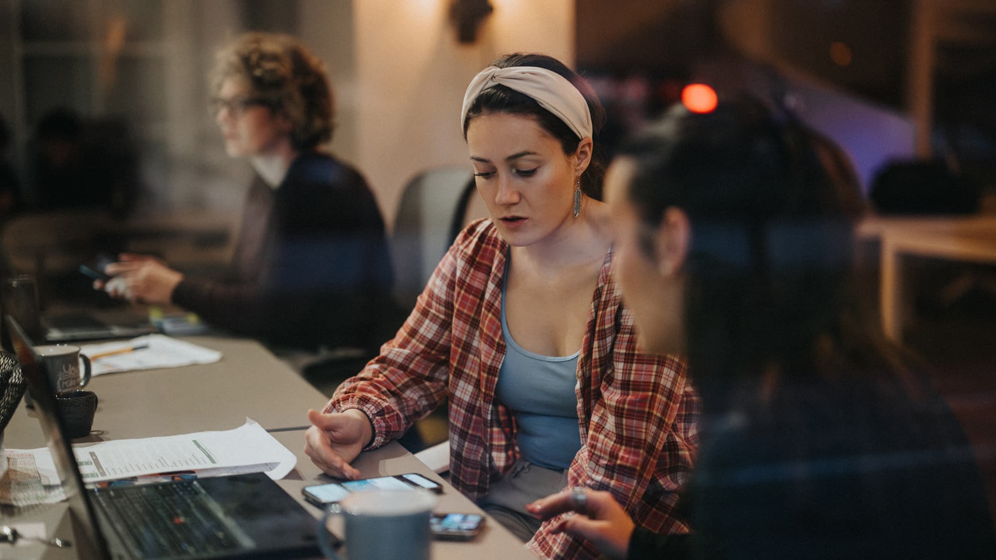 Zwei weibliche Kolleg+innen sprechen an einem Büro-Arbeitsplatz über ein Thema und schauen dabei auf technische Smartphones.