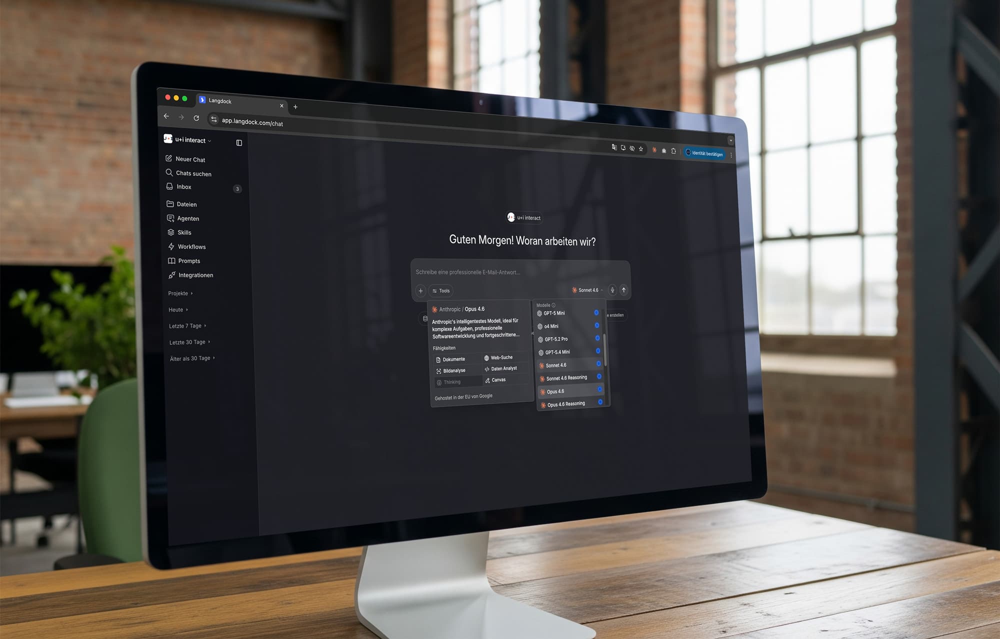 Ein großer Monitor steht auf einem Holztisch in einem modernen Büro mit Backsteinwänden. Auf dem Bildschirm ist die u+i interact KI-Plattform auf Langdock geöffnet. Zu sehen ist die Modellauswahl mit verschiedenen KI-Modellen wie GPT-5 Mini, Sonnet 4.6 und Opus 4.6 sowie eine Detailansicht des Modells „Anthropic Opus 4.6".