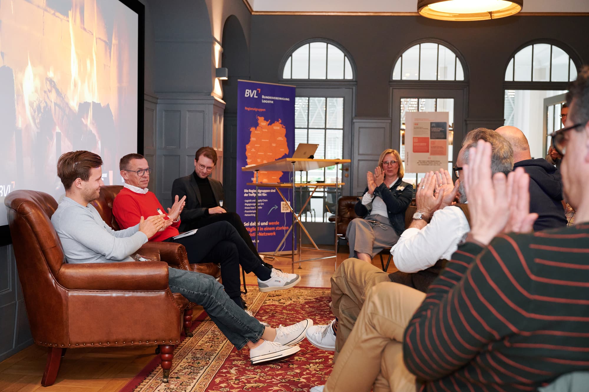 Gruppenfoto einer Gesprächsrunde in einem Raum mit hohen Rundbogen-Fenstern; die Gastredner des Abends sitzen vorne auf Ledersitzen. Das Publikum klatscht Beifall.