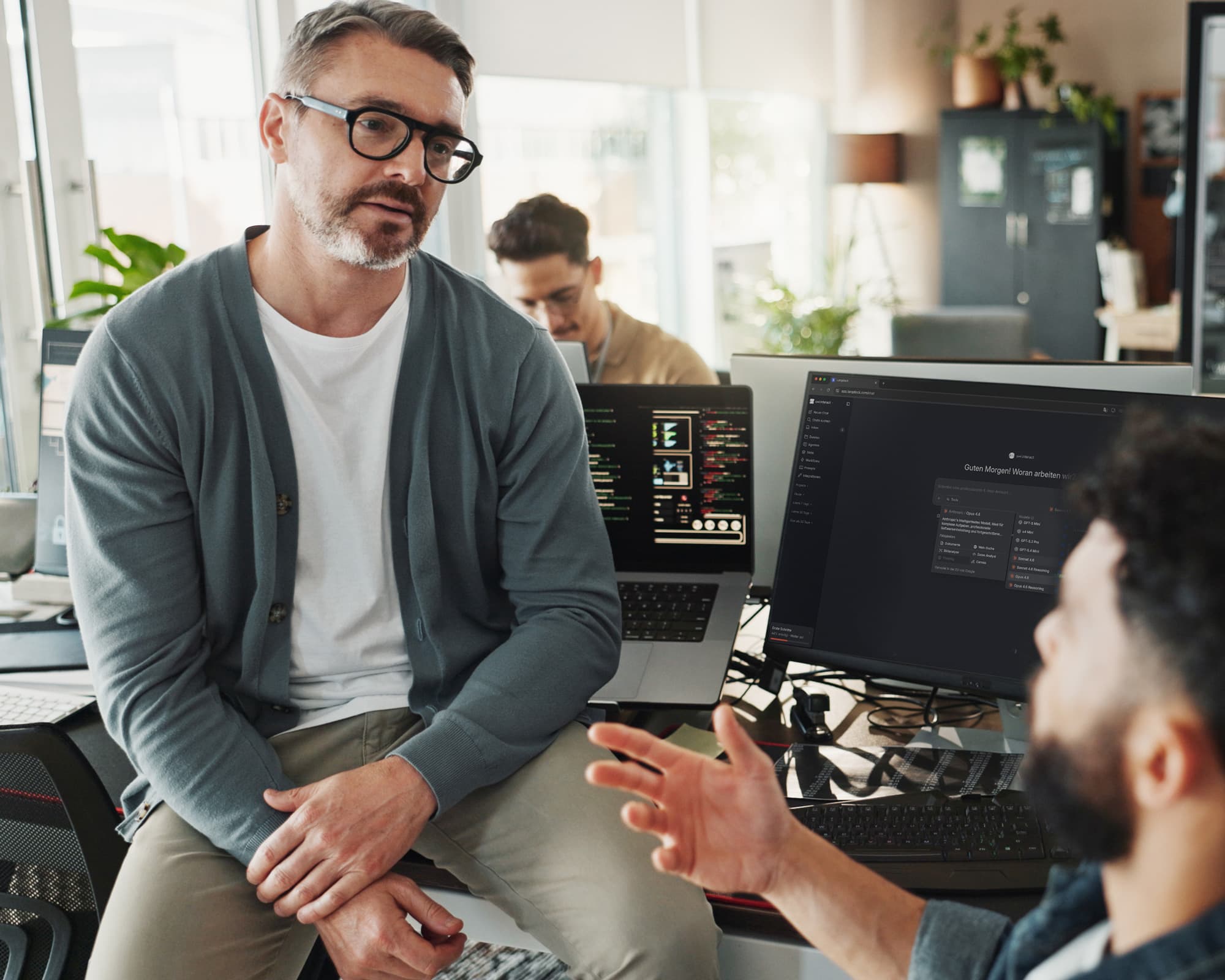 Zwei Kolleg+innen tauschen sich in einem Büro aus. Ein Mann mit Brille hört aufmerksam zu, während sein Gesprächspartner gestikulierend erklärt. Auf dem Monitor im Vordergrund ist die u+i interact KI-Plattform auf Langdock geöffnet. Im Hintergrund arbeitet eine weitere Person an einem Laptop.