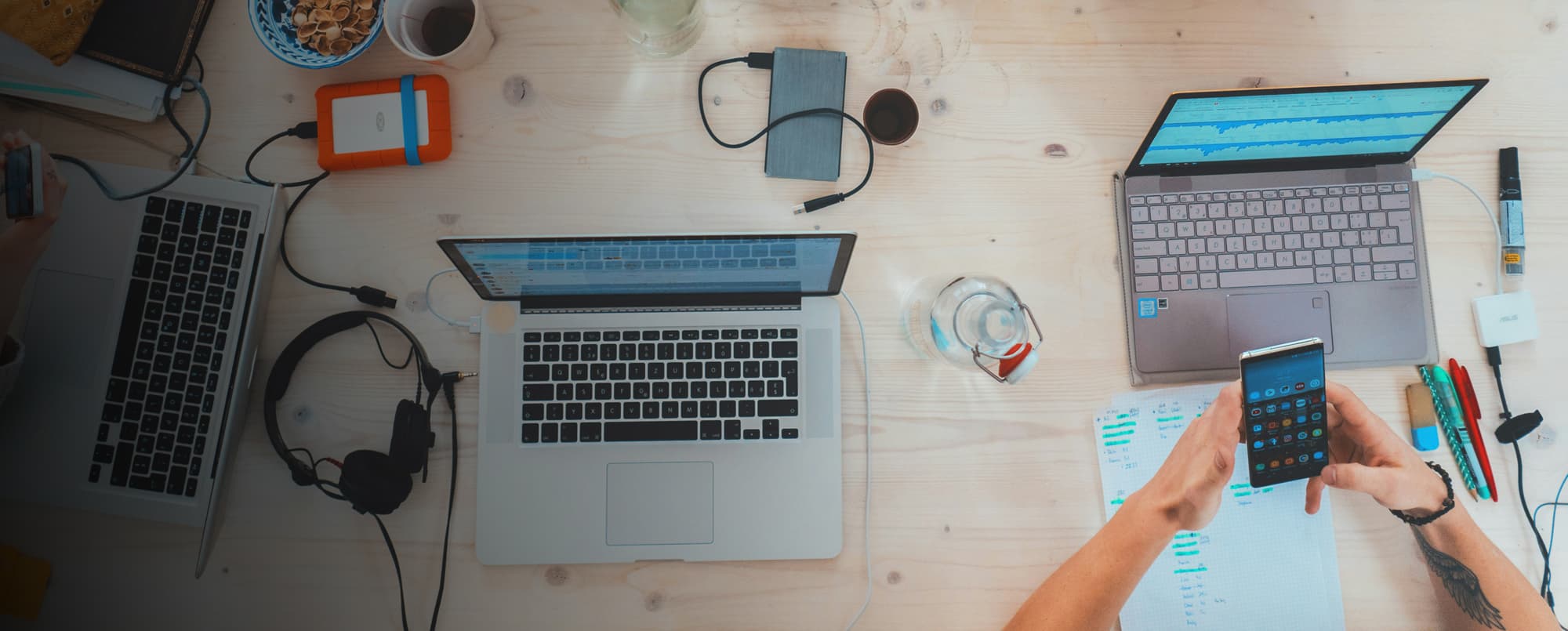 Oberansicht eines Holztisches mit mehreren Laptops, Smartphones und Notizbüchern, an dem mehrere Personen gemeinsam arbeiten. Digitale Geräte, Kopfhörer, externe Festplatten und Schreibutensilien sind verteilt, während Hände auf Tastaturen tippen oder ein Smartphone bedienen.