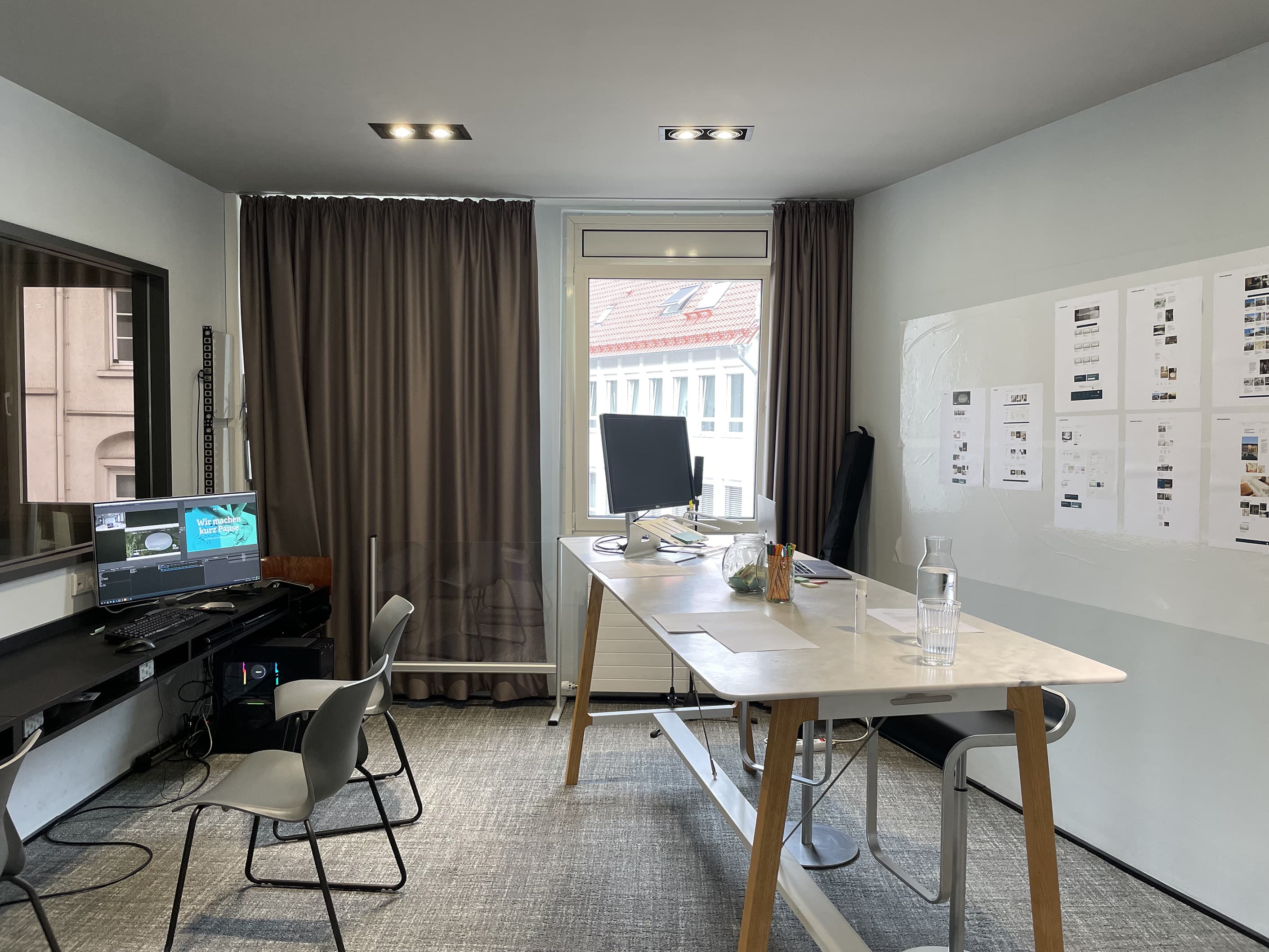 Blick auf einen Testraum in unserem Usability Teststudio, das an diesem Tag zum UX-Testing-Day für Interessierte geöffnet ist. An der Wand sind Layouts und Entwürfe angebracht. Ein Computerbildschirm zeigt eine Präsentation, während der Raum durch ein großes Fenster mit Blick auf benachbarte Gebäude beleuchtet wird.