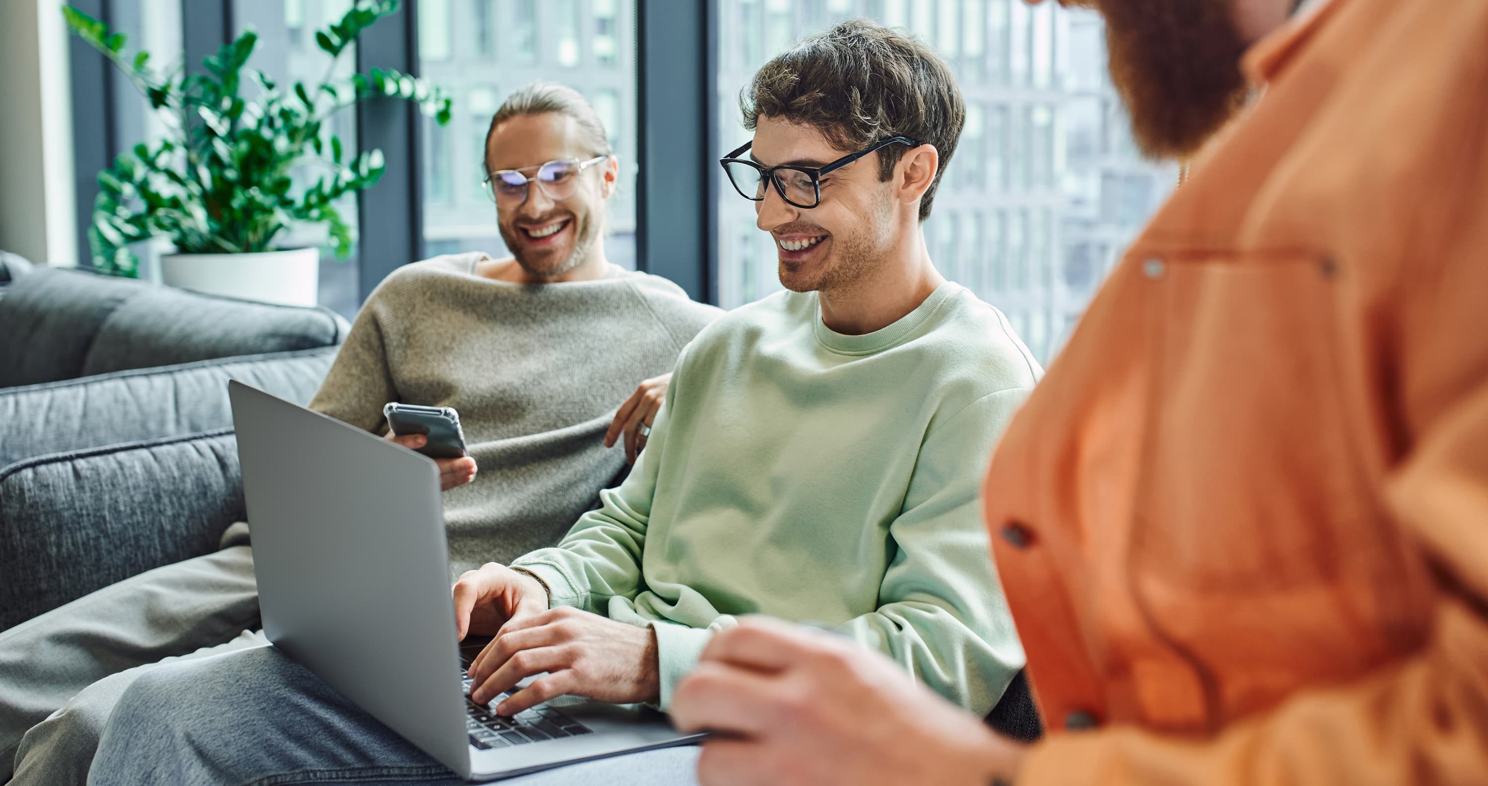 Drei junge Männer sitzen entspannt in einer modernen Büroumgebung mit großen Fenstern und Pflanzen im Hintergrund. Einer von ihnen arbeitet lächelnd an einem Laptop, während der Mann neben ihm, in einem grauen Pullover, ein Smartphone in der Hand hält und ebenfalls lächelt. Vom dritten Mann ist nur die orangefarbenen Jacke leicht verschwommen im Vordergrund zu sehen.