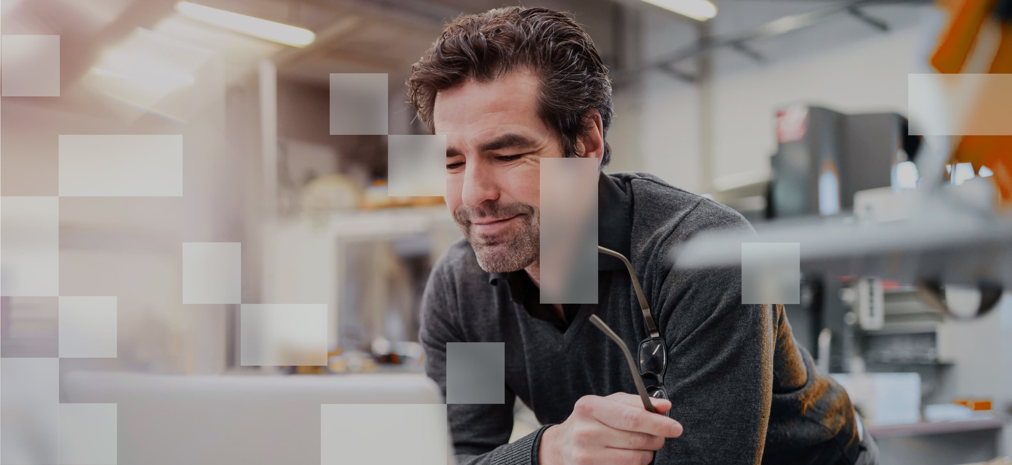 Ein Mann mittleren Alters mit dunklen Haaren und Bart lächelt leicht, während er sich über einen Schreibtisch beugt und eine Brille in der Hand hält. Im Hintergrund befindet sich eine moderne, industrielle Arbeitsumgebung. Transparente, weiße Quadrate überlagern das Bild, wodurch ein digitales und abstraktes Design entsteht.