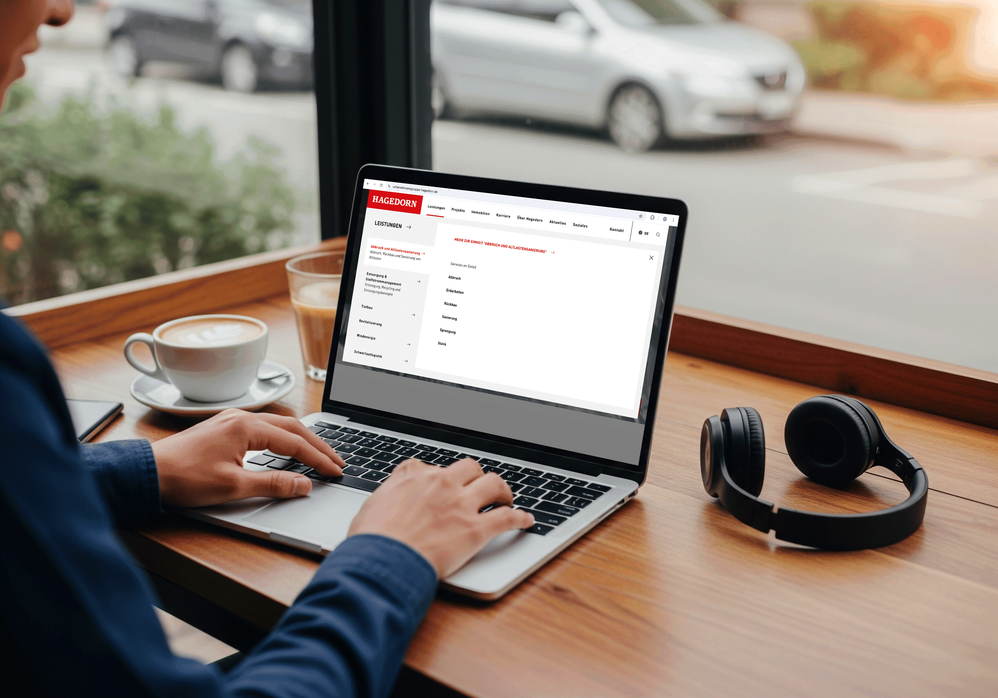 Person, die an einem Holztisch in der Nähe eines Fensters auf einem Laptop tippt, mit Kopfhörern, einem Smartphone und einer Tasse Kaffee in der Nähe. Draußen sind Autos zu sehen.