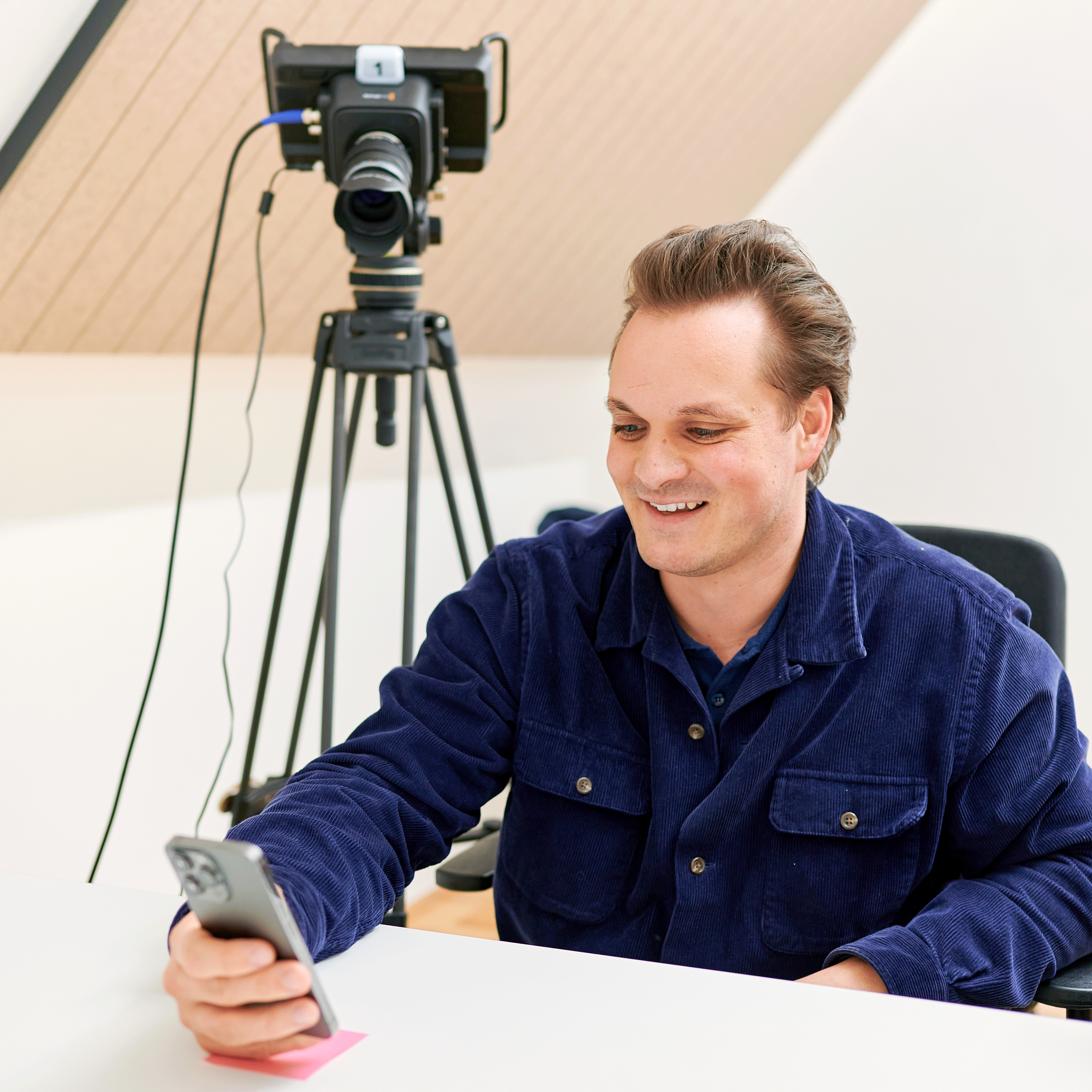 Ein lächelnder Mann in einer blauen Jacke, der an einem Schreibtisch sein Smartphone benutzt, im Hintergrund eine professionelle Kamera und ein Stativ.