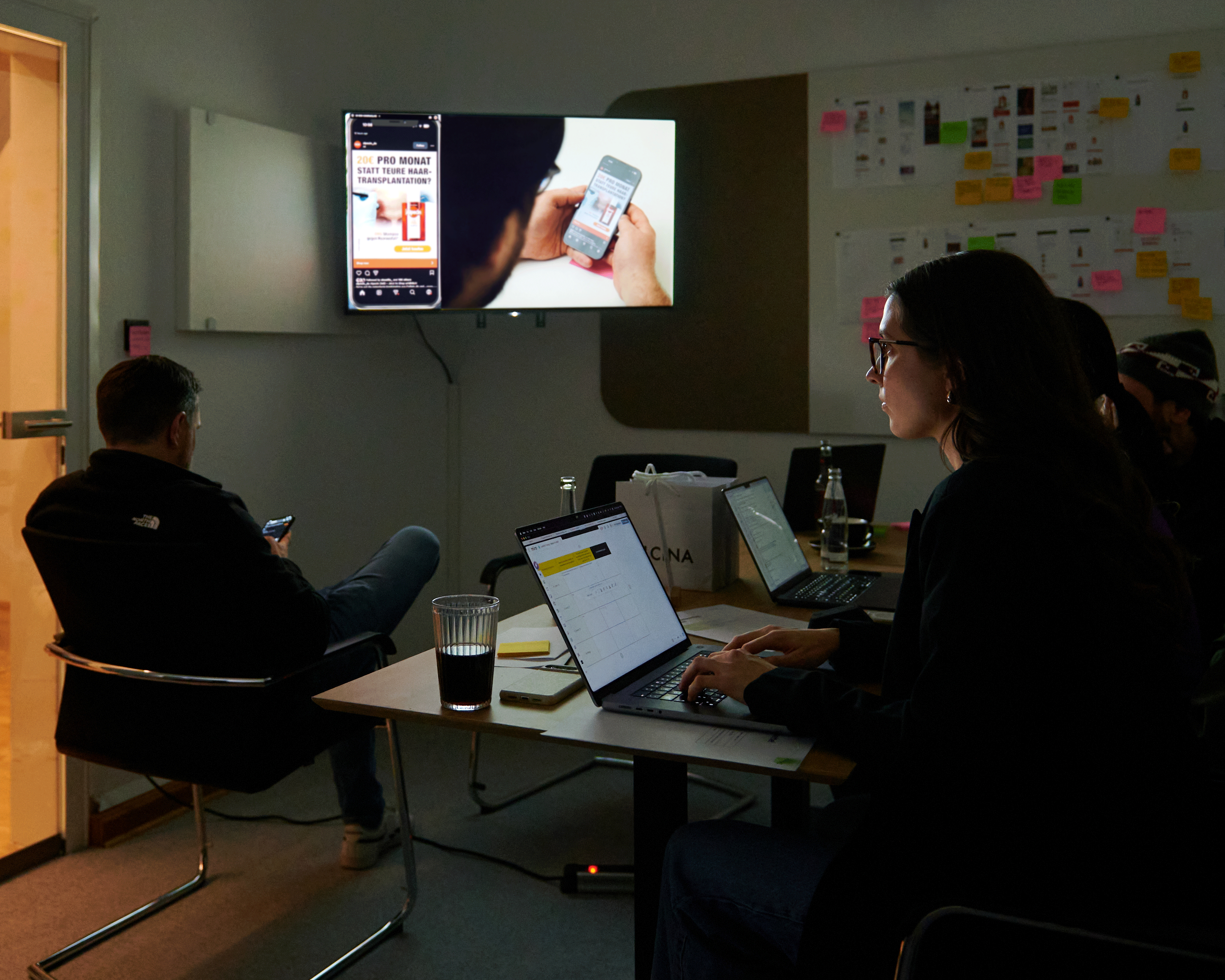 Eine Gruppe von Menschen in einem schwach beleuchteten Raum, die an Laptops arbeiten, auf deren großen Bildschirmen eine Smartphone-Oberfläche zu sehen ist.
