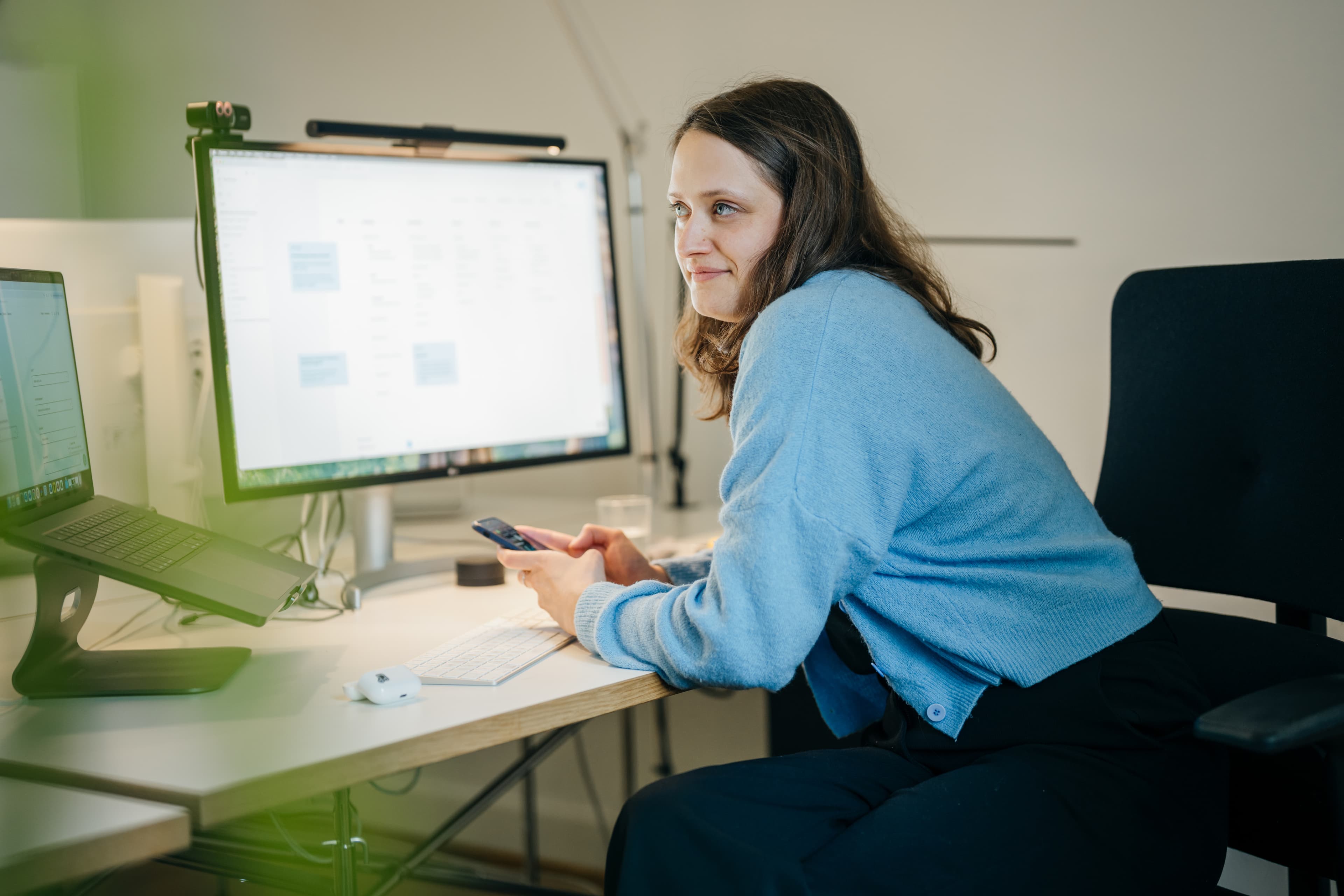 Eine Mitarbeiterin in einem blauen Pullover sitzt an einem Schreibtisch mit zwei Monitoren und hält ein Handy in der Hand.