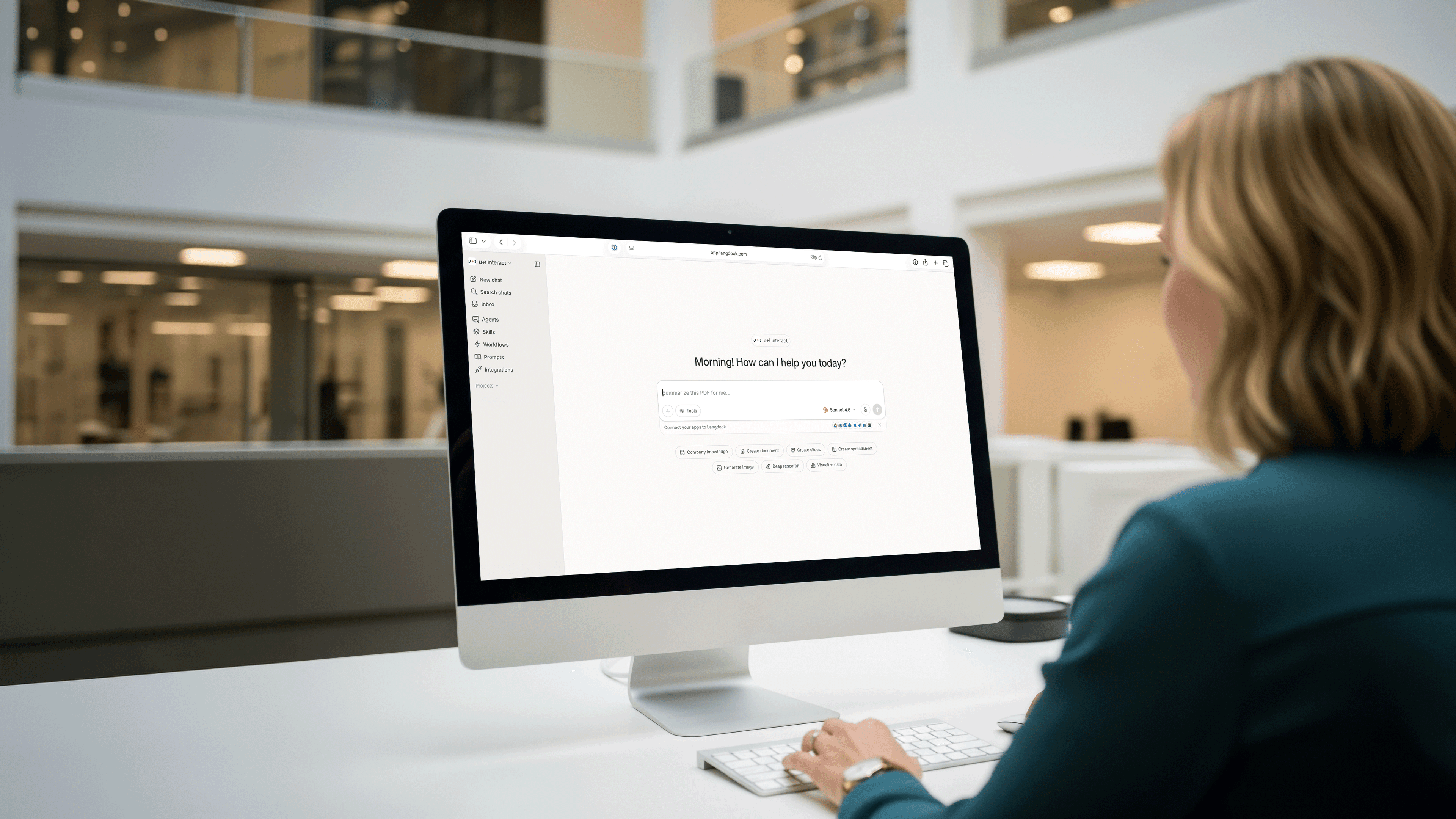 Eine Person in einem Büro sitzt an einem Schreibtisch vor einem verschwommenen Bürohintergrund und nutzt einen Computer, auf dem die Benutzeroberfläche des Langdock-Assistenten angezeigt wird.