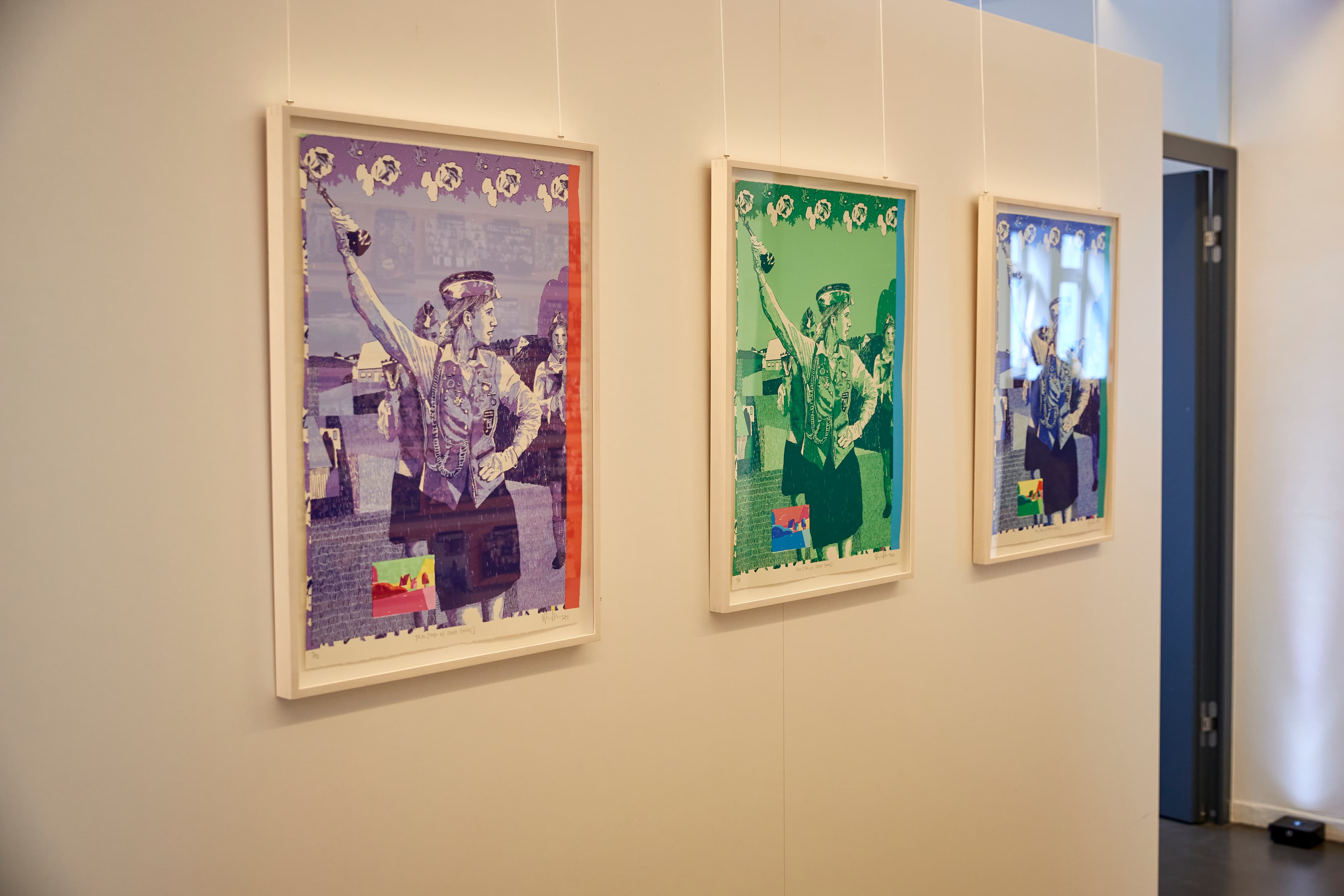 Drei gerahmte Kunstwerke, die nebeneinander an einer Wand hängen, zeigen jeweils eine Frau in Uniform, die eine Hand in die Luft hebt. Die Bilder sind in verschiedenen Farbschemata gehalten – lila, grün und blau – und haben einen collageartigen Stil. Die Ausstellung befindet sich in einem modernen Innenraum.