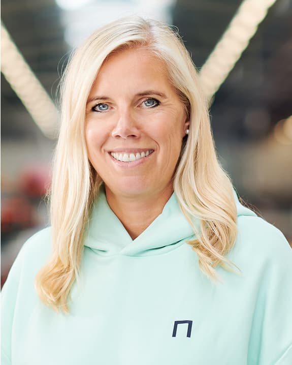 Smiling woman with long blonde hair wearing a light green hoodie, standing indoors with a blurred background.