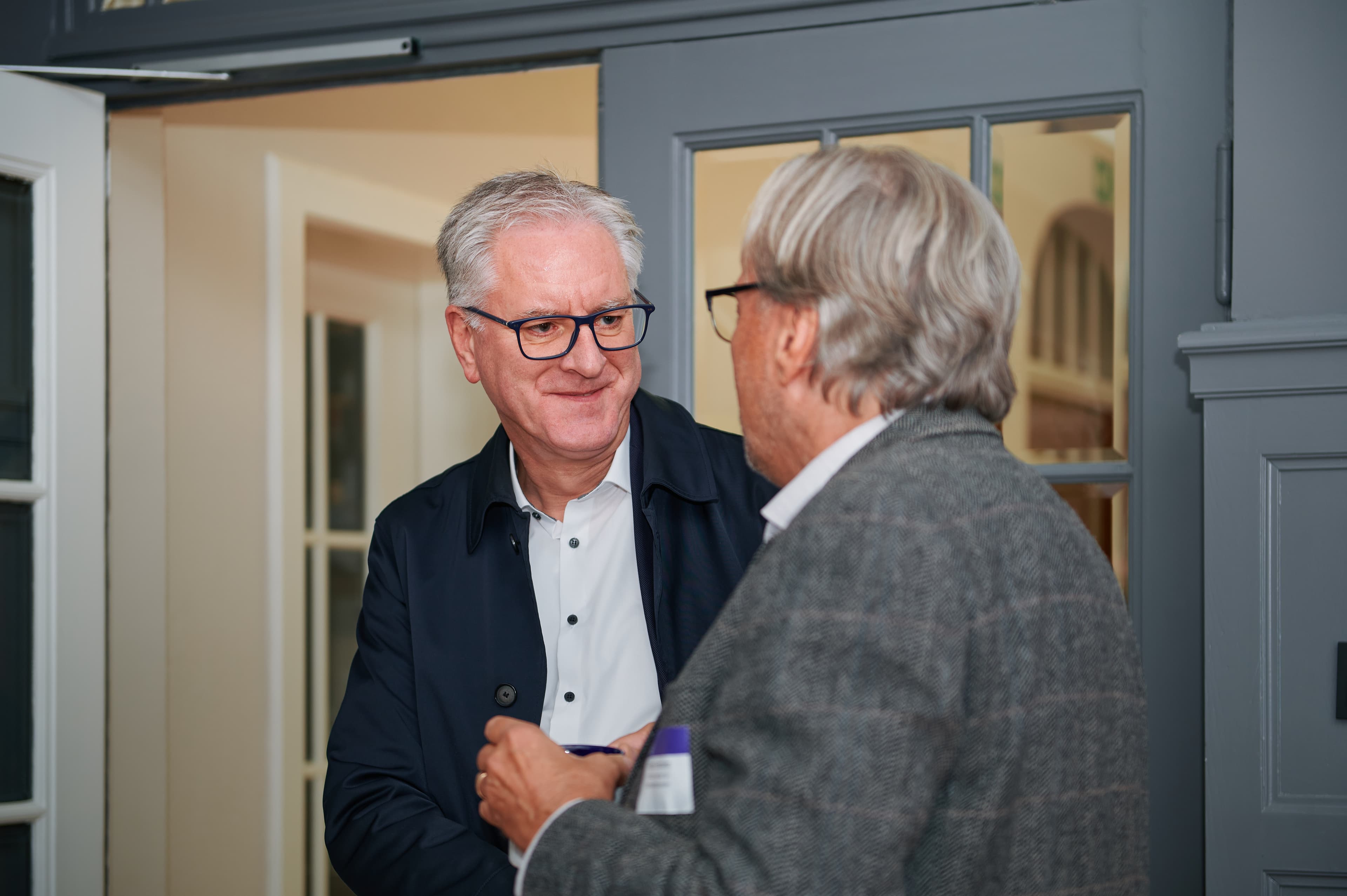 Two men in conversation near a doorway. One wears a dark jacket and white shirt, the other a gray blazer. Indoor setting with soft lighting.