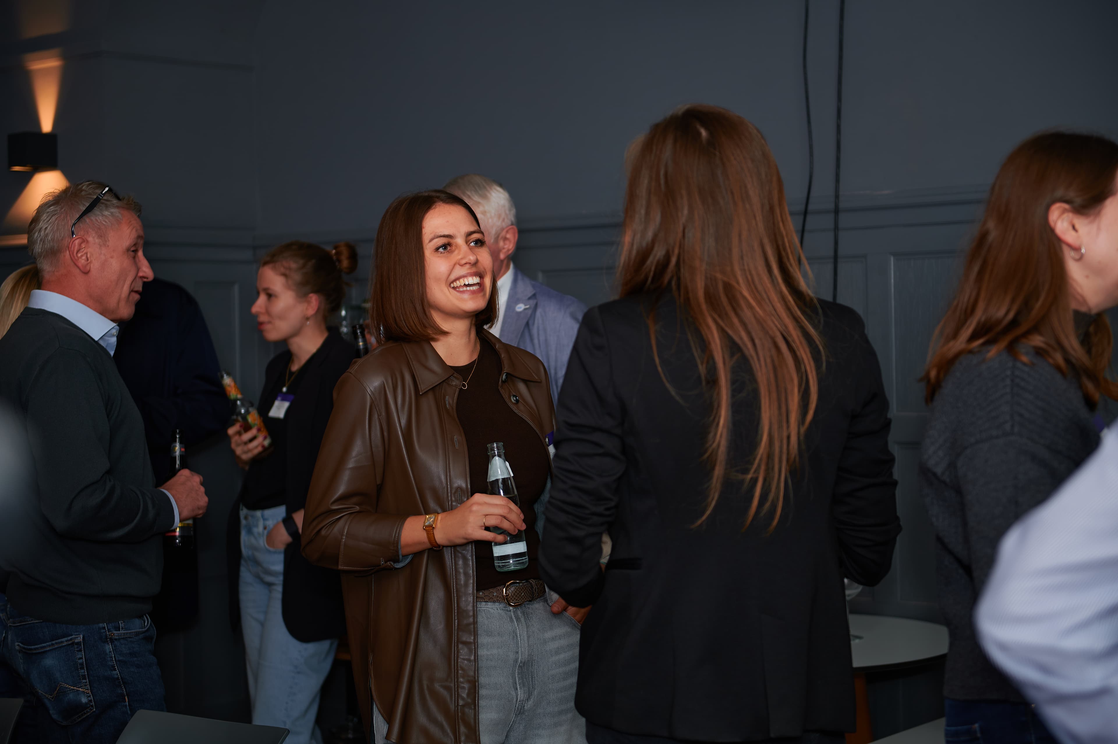 People socializing at an indoor event, with a woman in the foreground holding a drink and smiling, surrounded by others in casual attire.