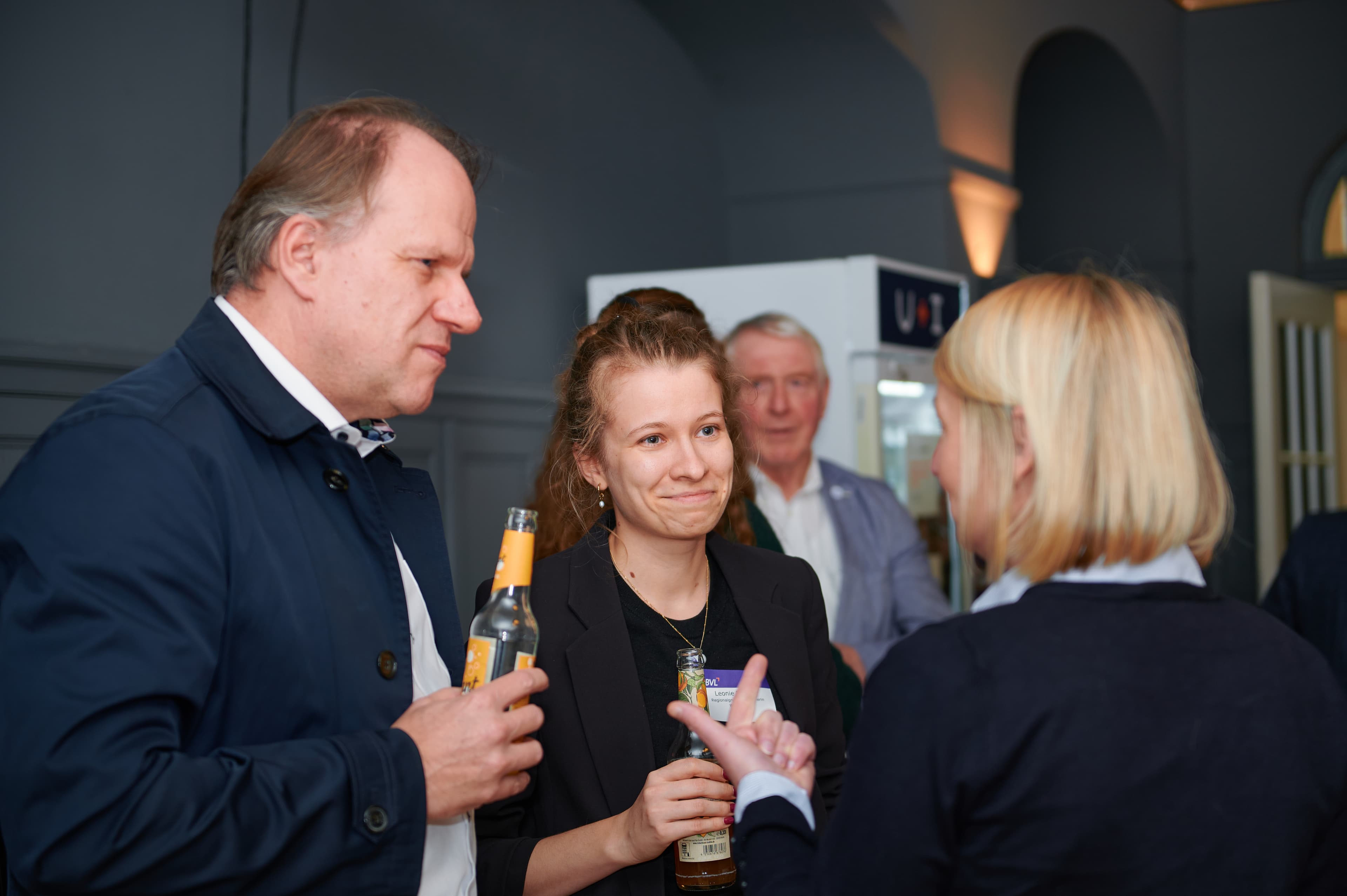 Three people are engaged in conversation at a social gathering, holding drinks, with a fourth person visible in the background.