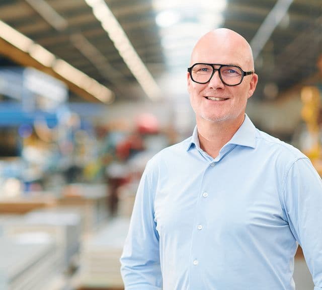 Smiling man in glasses and light blue shirt stands in a spacious, blurred industrial warehouse.