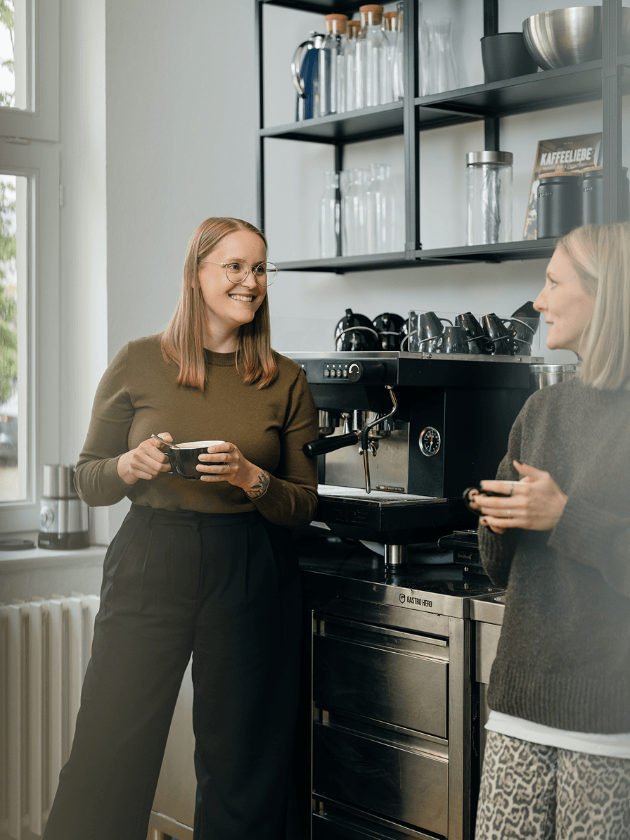 Zwei Frauen lächeln und unterhalten sich, während sie Kaffeetassen in den Händen halten. Sie stehen in einer modernen Küche mit einer Kaffeemaschine und Regalen im Hintergrund.