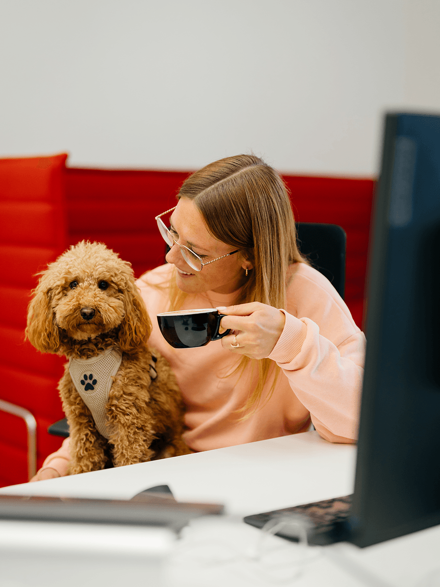 Eine Frau in einem rosafarbenen Pullover sitzt an einem Schreibtisch, hält eine Kaffeetasse in der Hand und hat einen kleinen braunen Hund auf dem Schoß, der ein Geschirr mit Pfotenabdruck trägt. Der Hund guckt direkt in die Kamera.